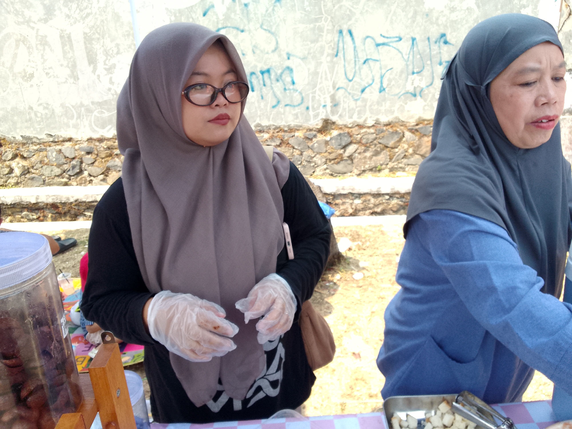 The image shows two women wearing hijabs at an outdoor market stall. The woman on the left is wearing a grey hijab and glasses, and appears to be attending to the stall. She is wearing clear plastic gloves. In front of her, to the left, is a large clear plastic container filled with dark, possibly dried, food items. To her right, a tan-colored bag is visible. The woman on the right is wearing a dark grey hijab and a blue top. She is looking down and to the right. In front of her, a small metal tray contains cubed white food items and a pair of tongs. The background consists of a textured wall with graffiti, a stone embankment, and dry, uneven ground. The overall scene suggests a food vendor setting in an outdoor environment.