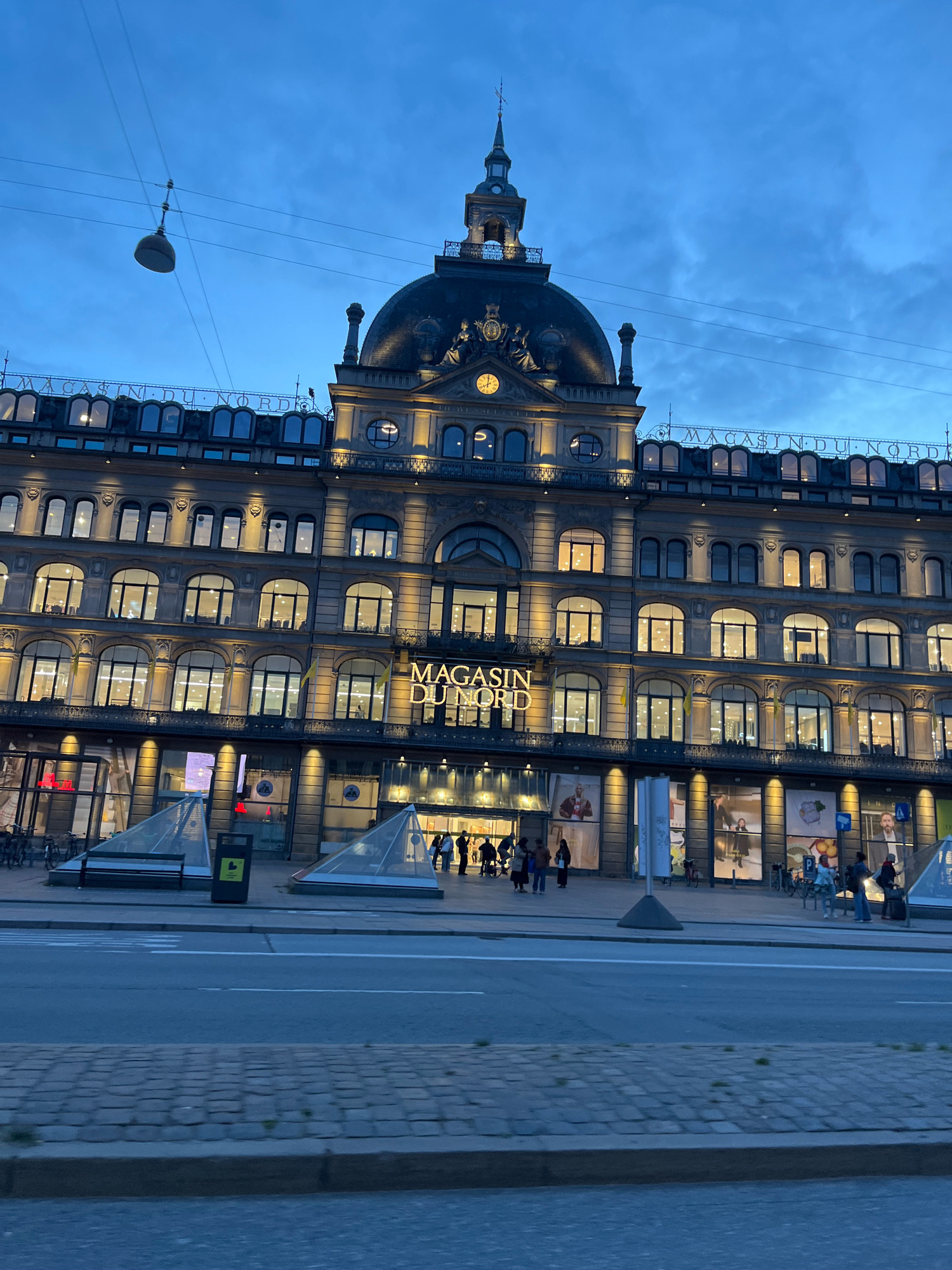 The image shows the Magasin du Nord department store in Copenhagen, Denmark at dusk. The building is grand and historic, with a prominent dome and clock tower. The facade is illuminated with warm lights, highlighting the many windows and architectural details. "MAGASIN DU NORD" is displayed in large, illuminated letters above the entrance. People are seen entering and exiting the store. In the foreground, a street with a pedestrian crossing and a cobblestone sidewalk is visible. Overhead, power lines and a street lamp are present against the twilight sky. The overall scene suggests a bustling urban environment as the day transitions to night.
