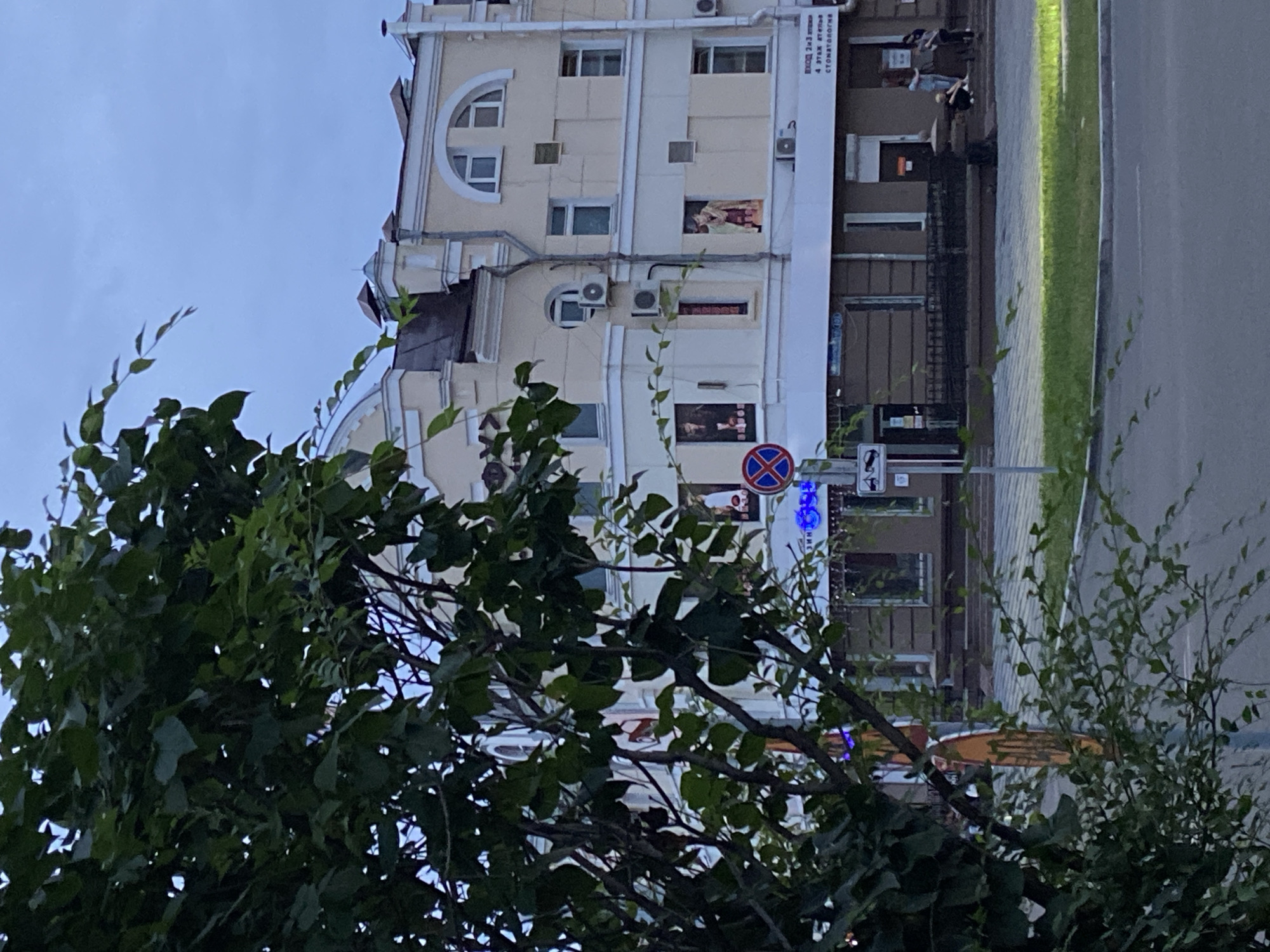 In Blagoveshchensk, Russia, this outdoor urban scene depicts a multi-story building with a light-colored facade, partially obscured by the branches and leaves of a large tree in the foreground. The building features multiple windows, some arched, and air conditioning units. The lower level of the building has a dark-colored storefront with visible signage and a few people standing outside. A traffic sign with a blue circle and a red cross is visible on a pole to the left of the building. The sky is overcast, and the lighting suggests it is either early morning or late afternoon.