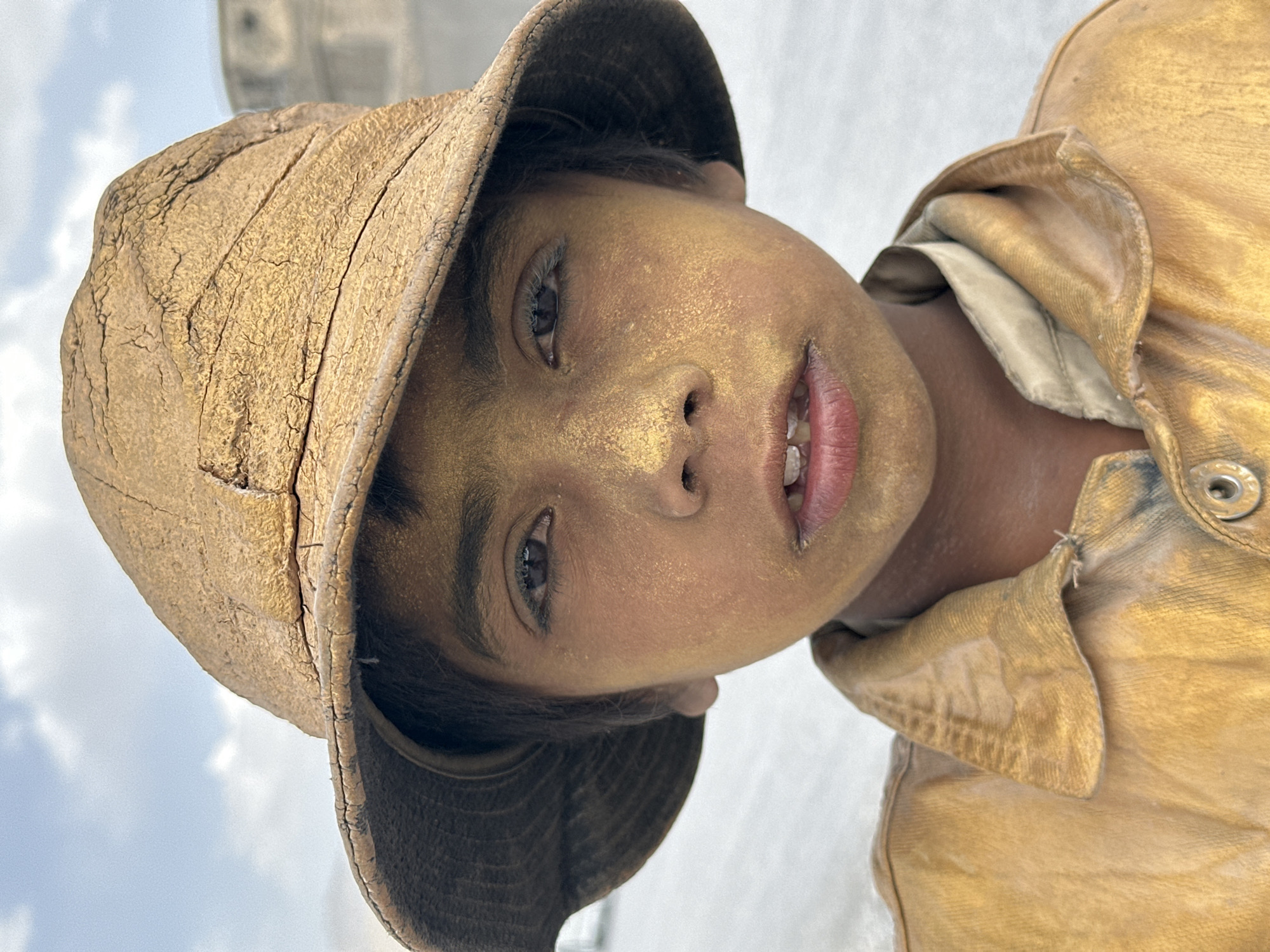 A young person with gold-tinged skin and wearing a worn, brown hat and yellowish-brown shirt looks directly at the camera. The image appears to have been taken outdoors, with a glimpse of sky and possibly buildings in the background. The person's face has a dusting of gold-colored material. It is plausible that the location is Quetta City Tehsil, Pakistan, based on the photo's origin.