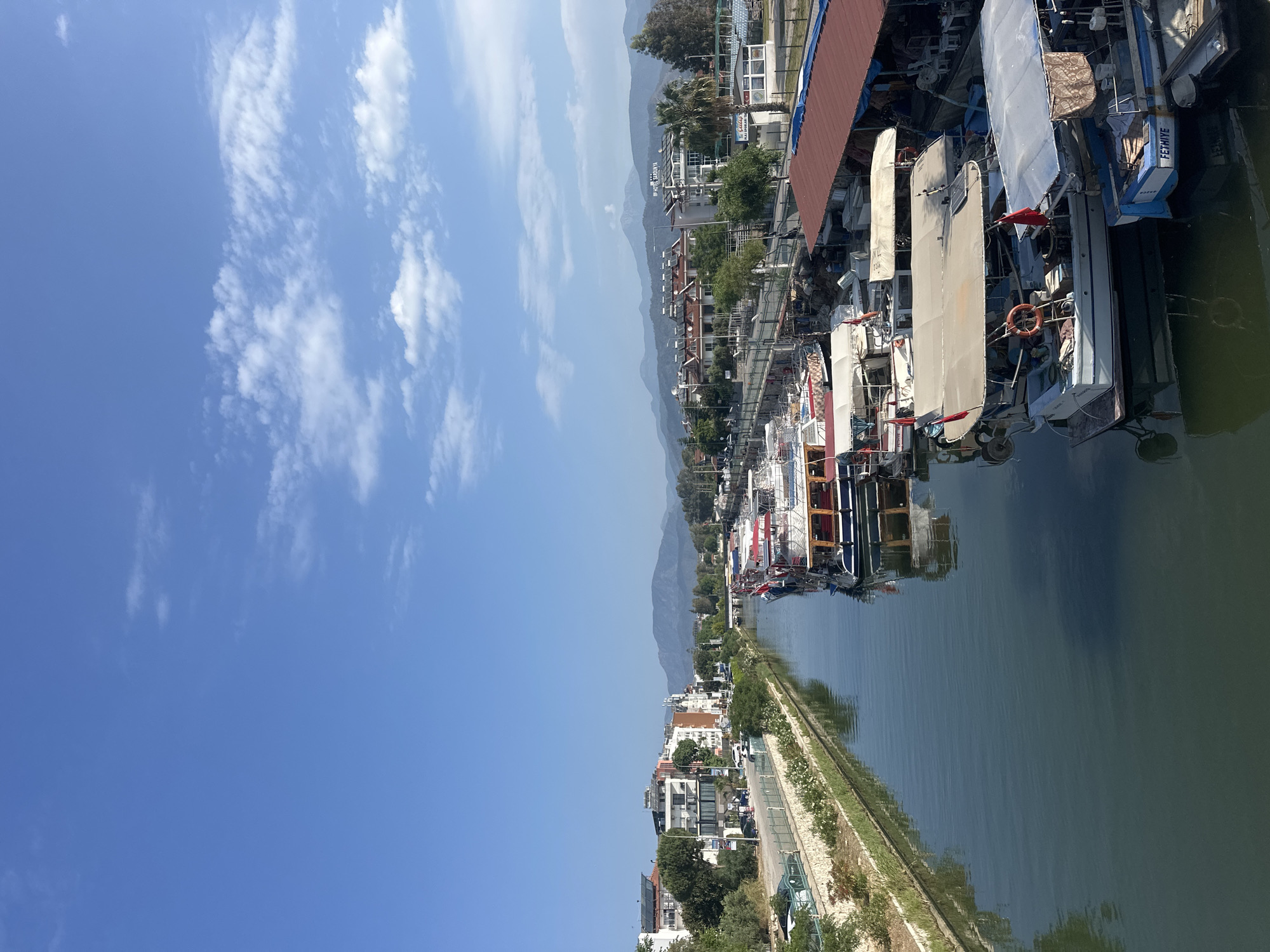 The image depicts a waterfront scene in Fethiye, Turkey, featuring a canal lined with boats. The boats are docked along a pier or quay, some appearing to be tour boats or fishing vessels. Buildings are visible along the canal, with a mountain range in the background. The sky is blue with scattered clouds. A road runs parallel to the canal on one side, with foliage and vegetation separating it from the water.