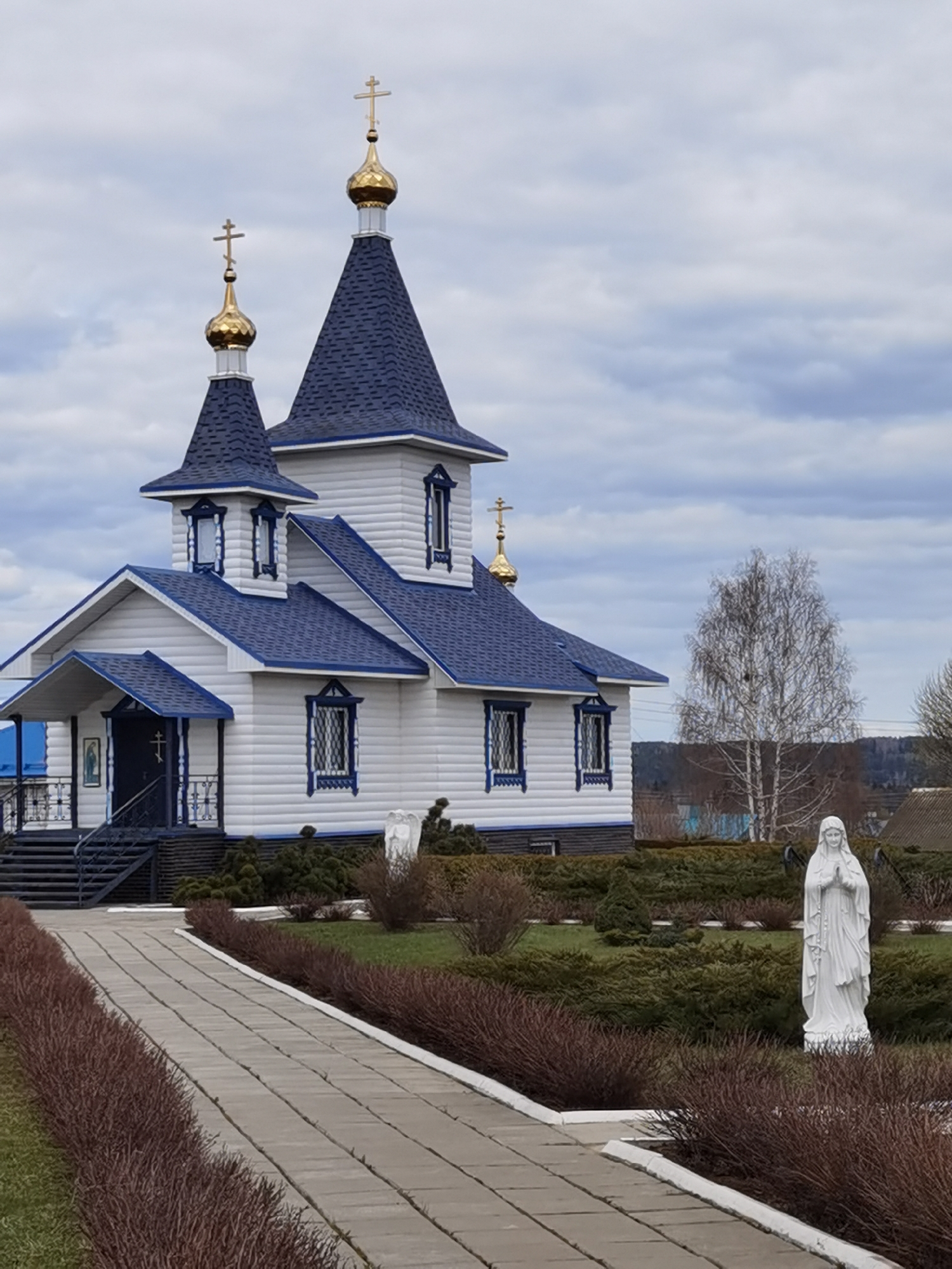 The image depicts a white church with blue accents, located in Кварса, Russia. The church has three golden domes topped with crosses. A statue of the Virgin Mary stands in the garden in front of the church, along a pathway. The sky is overcast. The surrounding landscape includes low shrubs and a bare tree.
