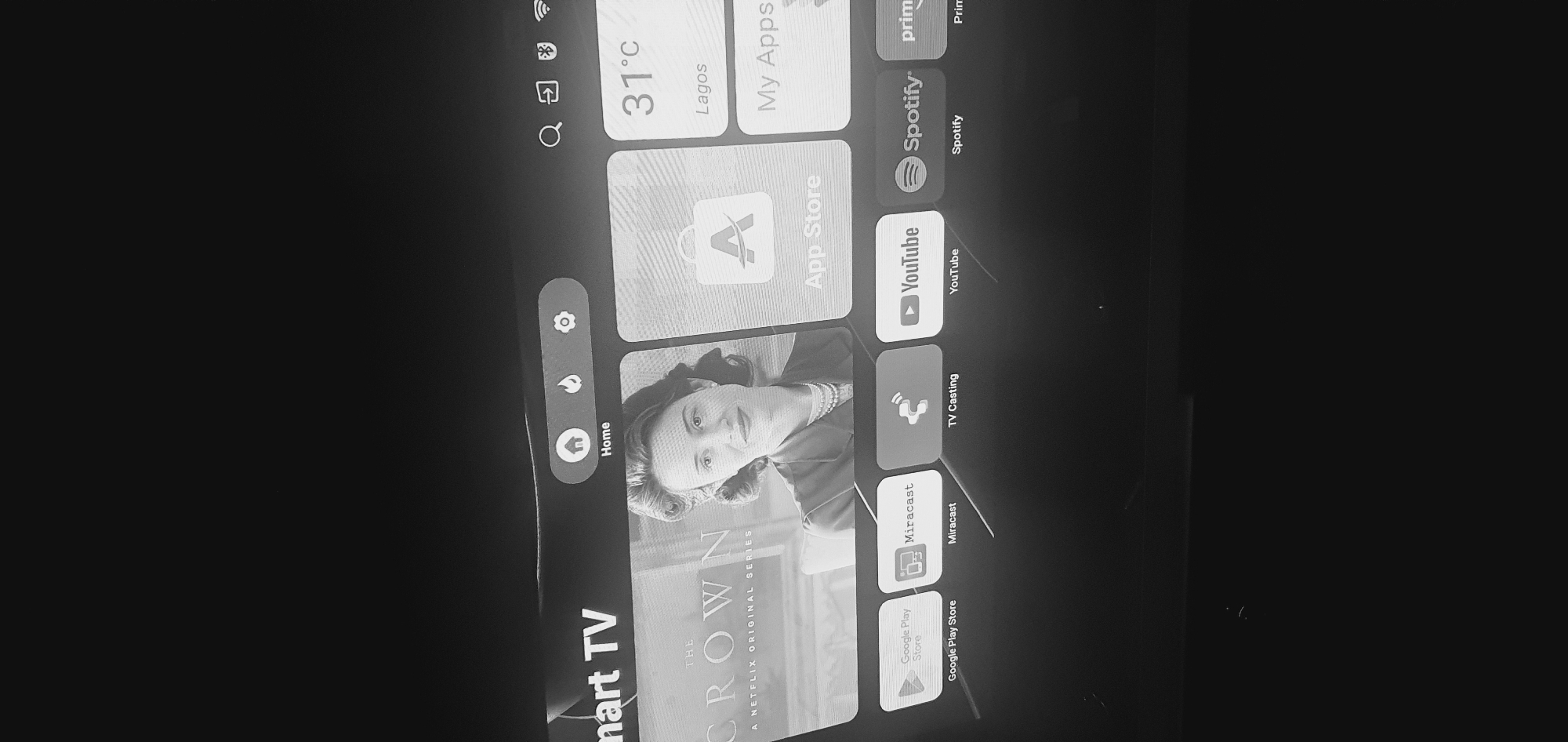 Here is a description of the image. The image shows a smart TV displaying a home screen with various applications. The TV screen features several app icons including Netflix's "The Crown", App Store, YouTube, Spotify, Google Play Store, and Miracast. Weather information indicates a temperature of 31°C in Lagos. The surrounding environment is dark, focusing attention on the illuminated TV screen. The image does not depict Lokoja, Nigeria, or any specific details about that location.