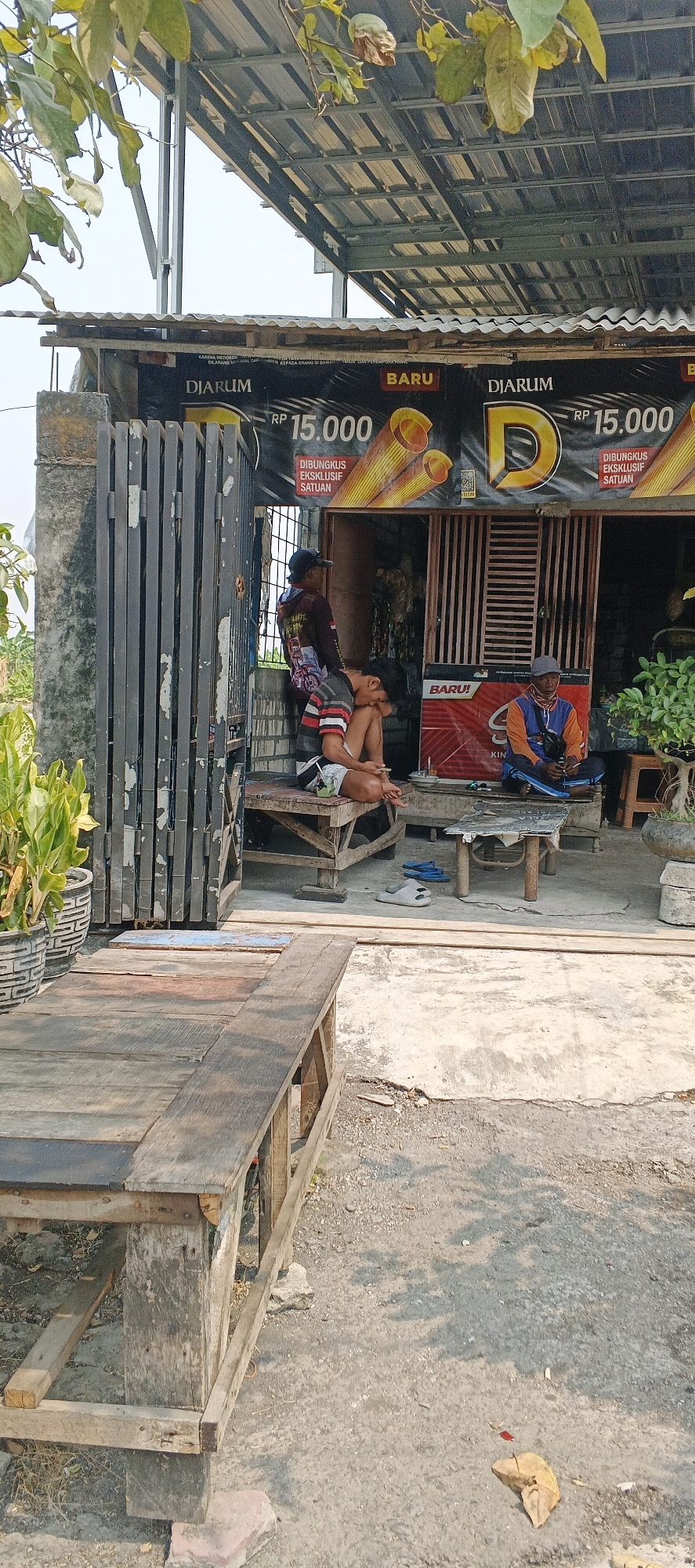 The image depicts an outdoor scene in front of a small shop or stall in Sonoadi, Indonesia. The focal point is a group of people relaxing in front of a building with a corrugated metal roof supported by a metal frame. A large banner advertising "DJARUM BARU RP 15.000" is visible on the building.
There are three men visible. One man is seated on a wooden bench, wearing a grey cap and an orange and blue shirt, holding a phone. Another man is seated on a lower wooden platform, wearing a black cap and a dark long-sleeved shirt with a colorful graphic. He is leaning forward with his head down. A third man, partially visible behind the gate, is wearing a dark shirt.
In the foreground, a rustic wooden table or bench made of planks is positioned diagonally. The ground is a mix of concrete and gravel. To the left, large potted plants with green leaves are situated near a weathered metal gate. The overall impression is a casual, everyday setting, possibly a local gathering spot or a shop in a rural or semi-urban area. The lighting suggests it is daytime, with shadows cast by overhead foliage.