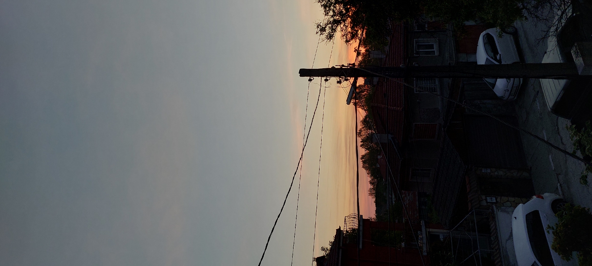 A low-angle, landscape-oriented shot displays a residential area at dusk in Yagodovo, Bulgaria. The sky blends from a muted grey to a band of orange and pink hues above the roofs of closely packed buildings. A wooden utility pole with electrical wires intersects the sky. Buildings with dark grey walls and red tile roofs occupy the right side of the frame. Windows are visible in the building facades. Parked cars are located on the street alongside the buildings. Trees obscure some of the rooftops. The photograph has been rotated 90 degrees clockwise.