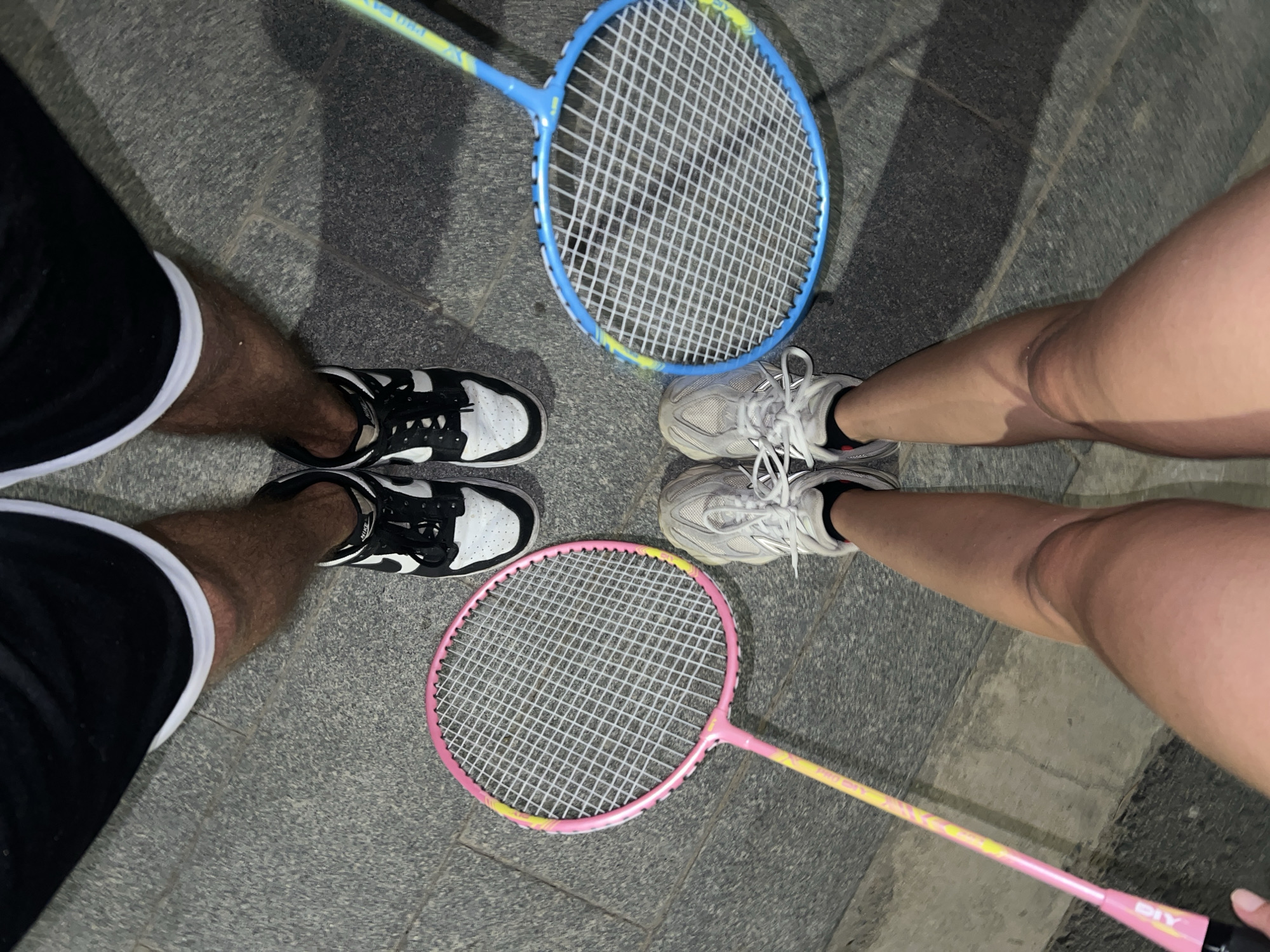 The image, captured from an overhead perspective, shows the feet and lower legs of two individuals positioned on a grey paved surface, possibly in Nam Nha Trang, Vietnam. Two badminton rackets are centrally placed between their feet.
On the left side of the frame, one person with darker skin is visible, wearing black shorts with a white stripe along the hem and black and white sneakers. On the right, another person with lighter skin wears light grey or white athletic sneakers.
A blue badminton racket, featuring a yellow gradient on its frame, rests between the left person's left foot and the right person's right foot. The text "PRO" is partially visible on the blue racket's shaft. A pink badminton racket, also with yellow accents on its frame, is positioned between the left person's right foot and the right person's left foot. The word "DIY" is clearly visible on the pink racket's handle.
The arrangement suggests the individuals are either preparing to play or have just finished a game of badminton. The lighting is dim, indicating it may be evening or night. The ground appears to be made of large, irregular grey paving stones or concrete tiles.