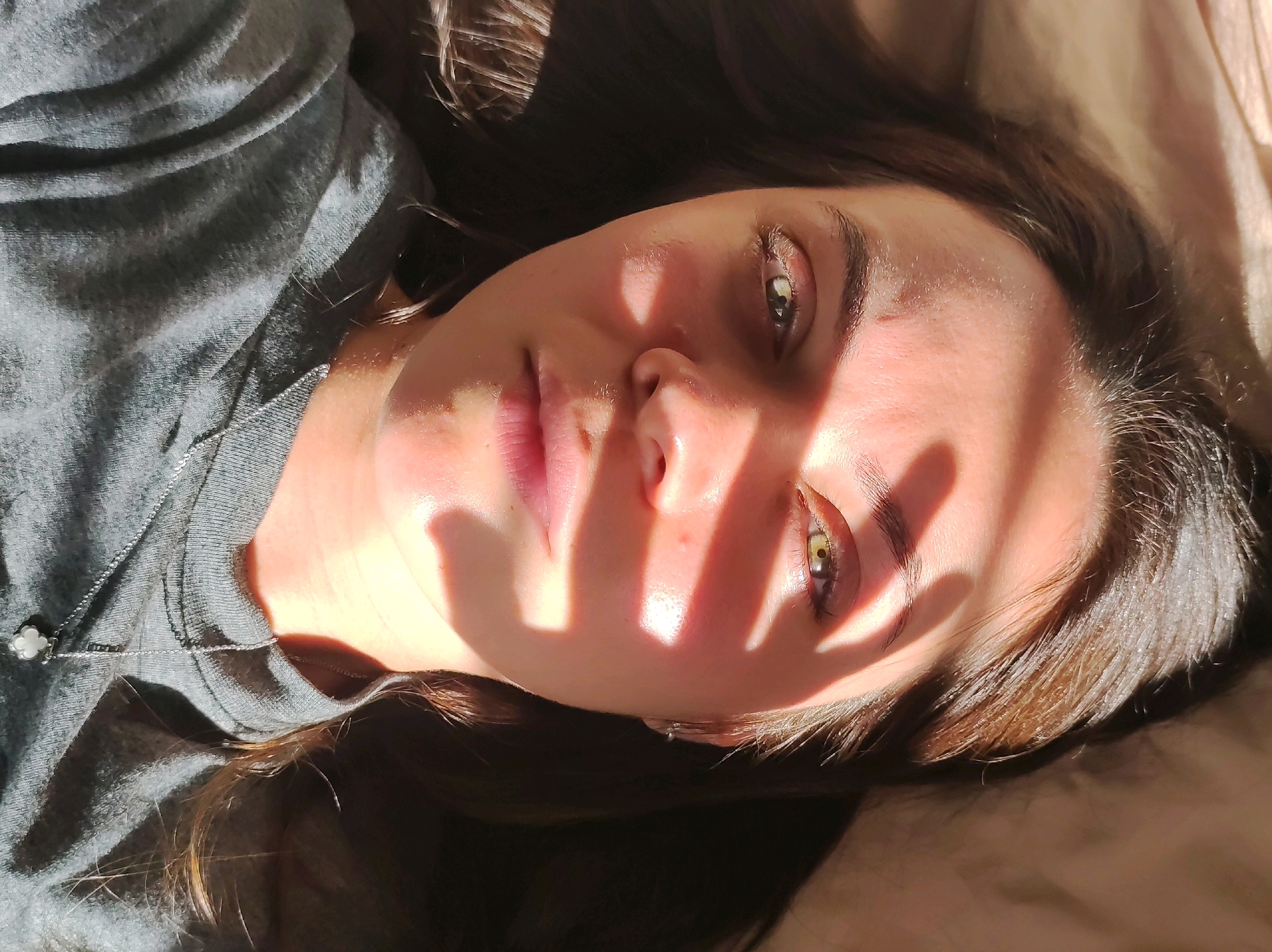 The image shows a close-up, overhead view of a young woman lying on her back, looking upwards. She is wearing a grey t-shirt and a delicate silver necklace with a white clover-shaped pendant. Sunlight streams across her face, casting a shadow of what appears to be window blinds or a similar object over her eyes and forehead. Her hair is dark brown and falls around her head. The background appears to be a bed or couch with textured fabric. The image does not provide any specific information about the location in Chaikovsky, Russia.
