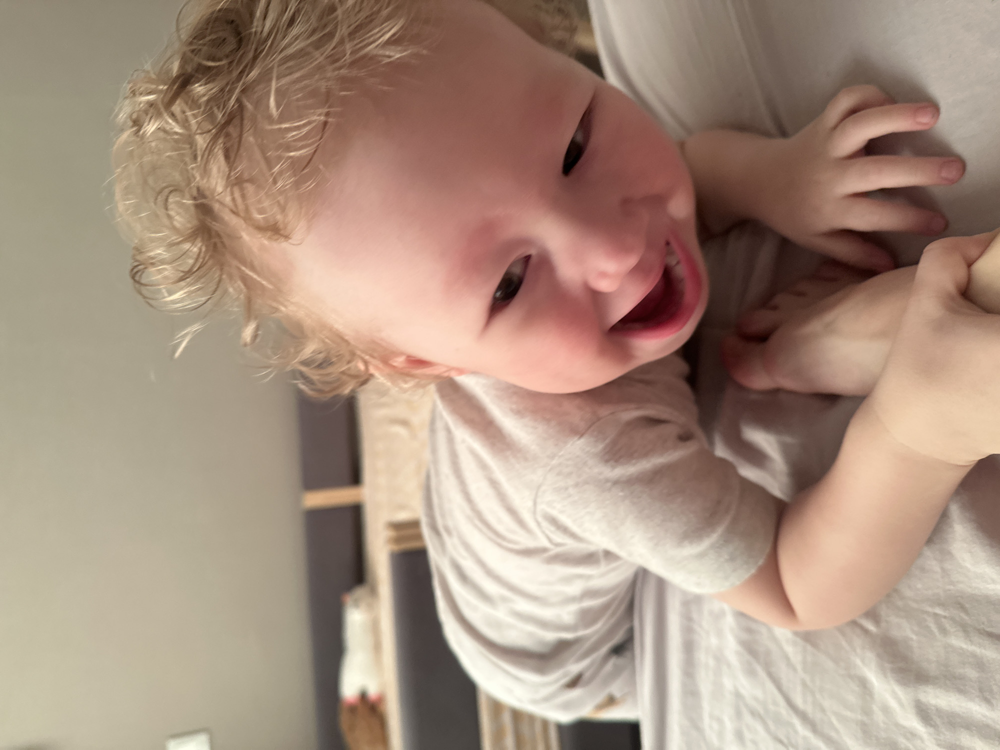 A child with light skin and curly blonde hair is being held in someone's arms, close to their chest. The child is wearing a light grey t-shirt and smiling. The person holding the child is wearing a light grey shirt. The background is a muted grey and includes part of a wooden structure and a white object. The location is unknown and cannot be determined as Votkinsk, Russia based on the visual data.