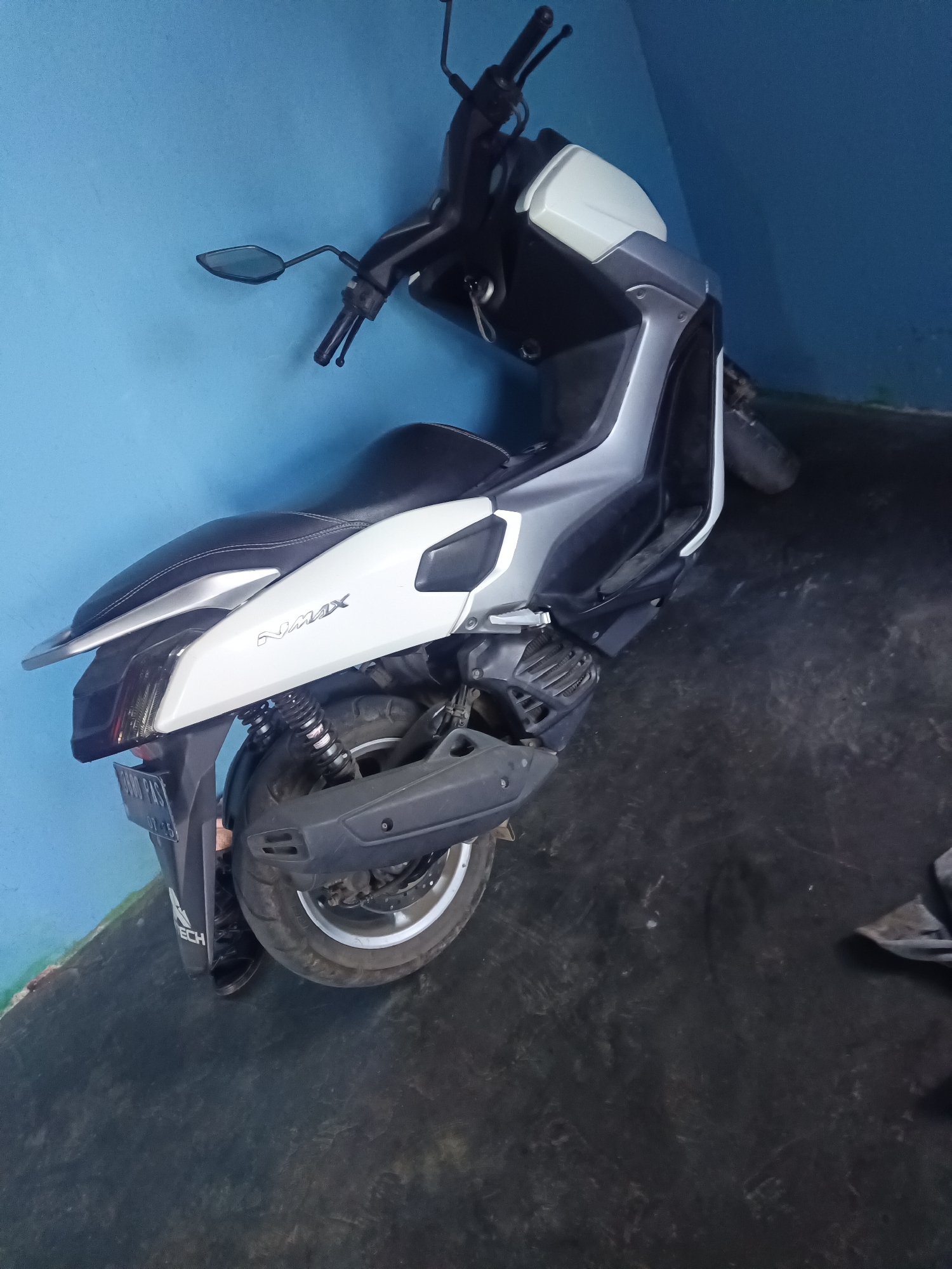 A white and grey Yamaha NMAX scooter is parked indoors, leaning against a bright blue wall. The scooter is positioned at an angle, with its front facing to the right and slightly upwards. The floor is a dark, textured surface, possibly concrete or asphalt, and appears somewhat worn. A license plate is partially visible on the rear fender, with some text and numbers discernible, although not entirely legible. The scooter's seat is black with white stitching. The background wall is a solid, vibrant blue. There are no people visible in the frame. The lighting suggests an indoor environment, possibly a garage or workshop. The image has a slight tilt to the left.