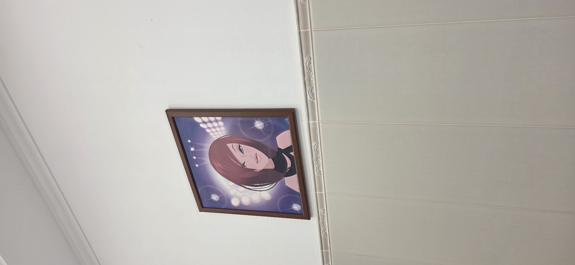 The image displays an indoor scene featuring a framed artwork mounted on a plain white wall. The artwork itself is a colorful illustration of a woman with reddish-brown hair, dark eyes, and full, slightly parted lips, depicted looking slightly upwards and to the right. She wears a dark top, possibly black, which exposes part of her shoulder. The background of the illustration is a vibrant purple and blue gradient, accentuated by multiple bright, circular spotlights or stage lights. A faint, illegible signature or text is visible in the upper right corner of the artwork.
To the right of the white wall, another wall or partition is visible. This surface is light grey or beige in color with a subtle textured pattern and is horizontally sectioned by thin, embossed lines. A decorative white molding runs vertically along the seam where this textured surface meets the plain white wall. The image provides a static view of interior decor, with no discernible activity taking place. The setting is an indoor space, located in Moscow, Russia.
