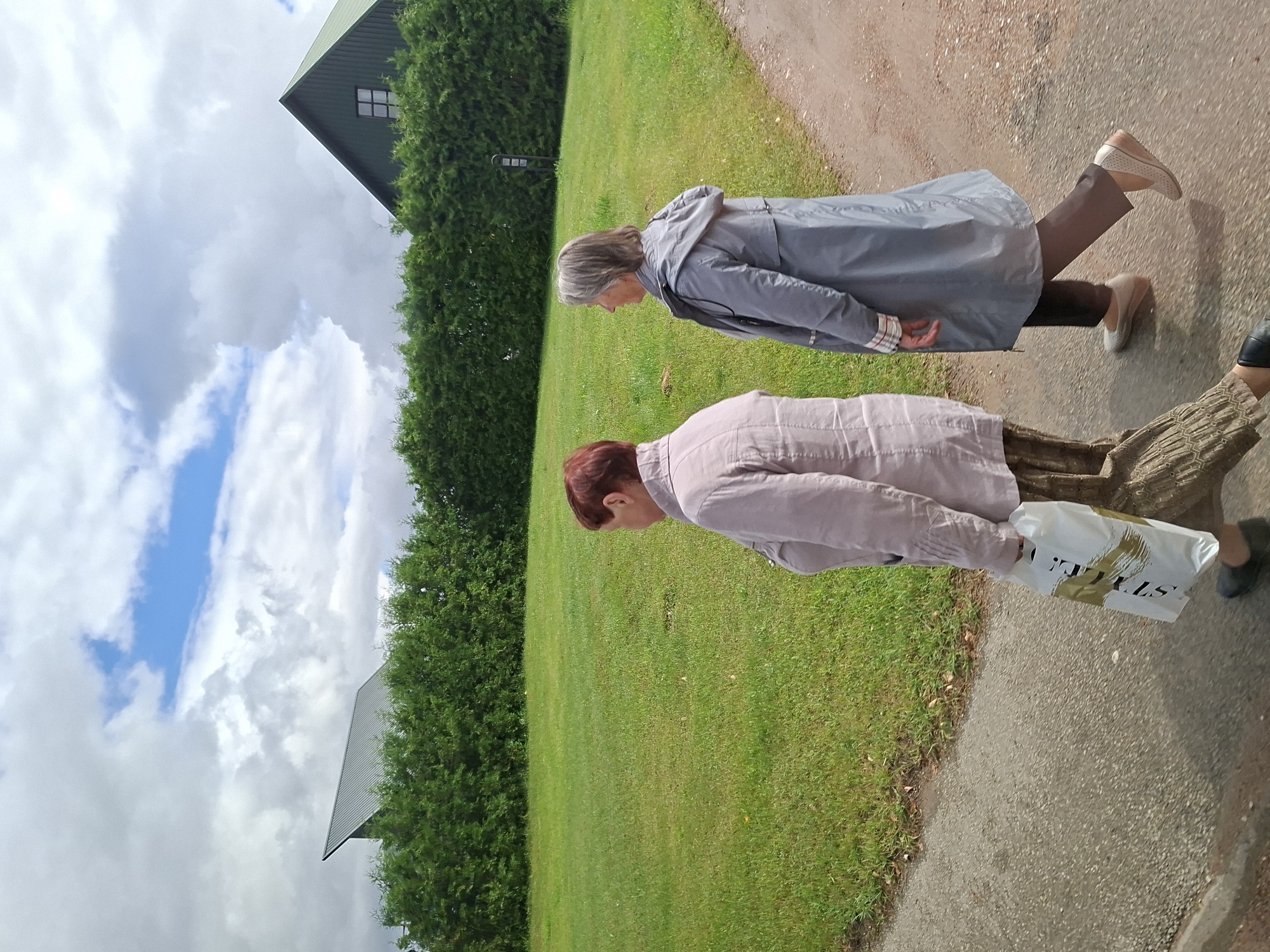 Two women are walking along a gravel path next to a grassy field in Balbieriškis, Lithuania. The woman on the left, with short red hair, is wearing a light-colored jacket and patterned pants and is carrying a white shopping bag. The woman on the right, with gray hair, is wearing a gray raincoat and dark pants. Behind them is a dense hedge and two buildings with dark green, sloping roofs, one of which has a multi-paned window. The sky above is partly cloudy with patches of blue.