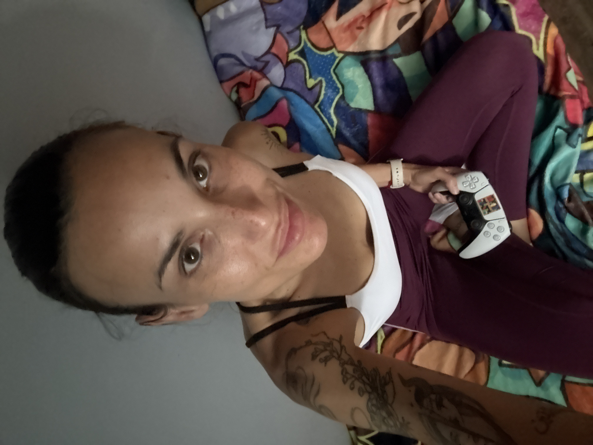 A young woman is depicted in a slightly angled, overhead selfie, relaxing indoors in Pleven, Bulgaria. She has dark hair pulled back from her face, warm brown eyes, and visible freckles, presenting a relaxed and friendly expression towards the camera. She wears a white sports bra or tank top with dark shoulder straps, paired with dark red or burgundy leggings. Her left arm is adorned with several tattoos, one clearly showing a detailed floral or vine-like design.
In her right hand, she holds a white PlayStation 5 DualSense controller, indicating engagement in video gaming. A light-colored wristband or smartwatch is visible on her right wrist. She is situated on a plain grey surface, likely a bed or floor, next to a vibrant, multi-colored blanket or duvet. This blanket features a striking pop art or comic book-inspired pattern with bold shapes in shades of purple, blue, green, orange, and black. The setting suggests a casual and comfortable home environment. No discernible text is visible on any objects.