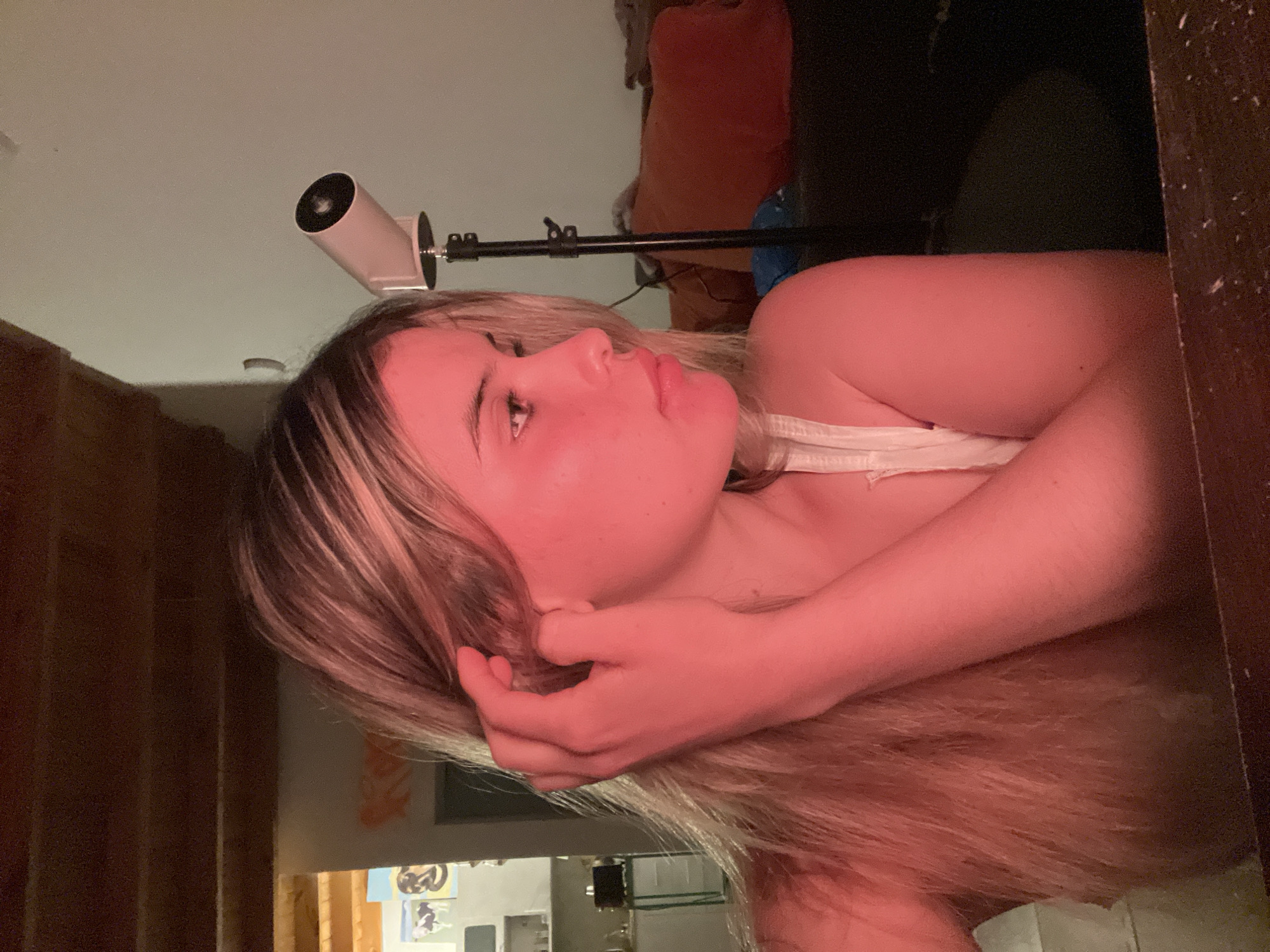 The image depicts a young woman with blonde hair, illuminated by a red light source, in a dimly lit interior setting. She is seen in profile, touching her hair. In the background, a tall lamp with a cylindrical head is visible, along with furniture including a couch with orange cushions. The environment appears to be a residential or studio space, with wooden beams and shelves visible. There is no indication of the location being in Sumalao, Argentina.