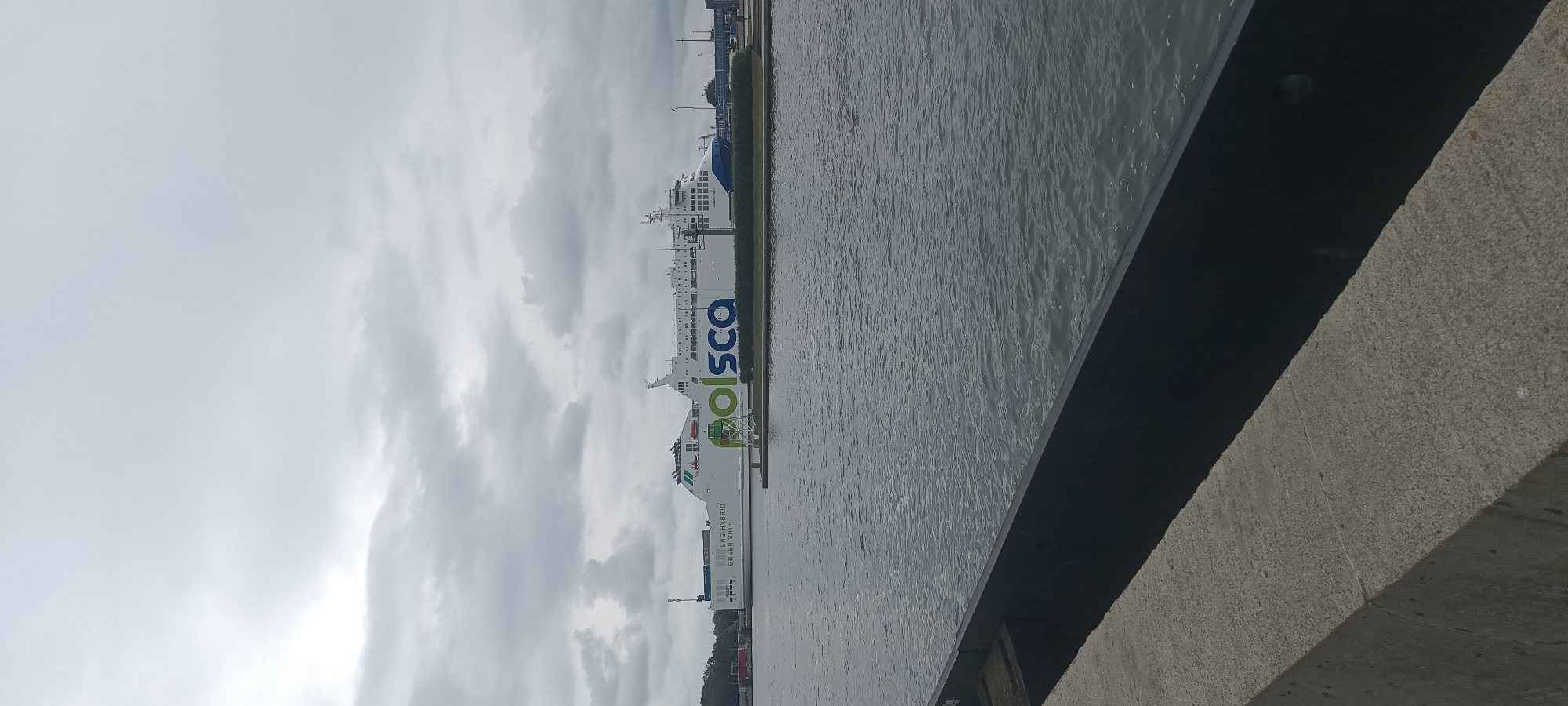 The image captures a wide view of a large, white ferry docked in a waterway. The ferry prominently displays the green and blue "polsca" logo. Text on the side of the ferry reads "LNG-HYBRID GREEN SHIP". The sky is overcast with clouds. In the foreground, a concrete embankment lines the water's edge. The location appears to be a port or harbor, consistent with the city of Świnoujście, Poland, known for its ferry terminal connecting to Scandinavia. No people are visible in the image. The water is calm, with subtle ripples.