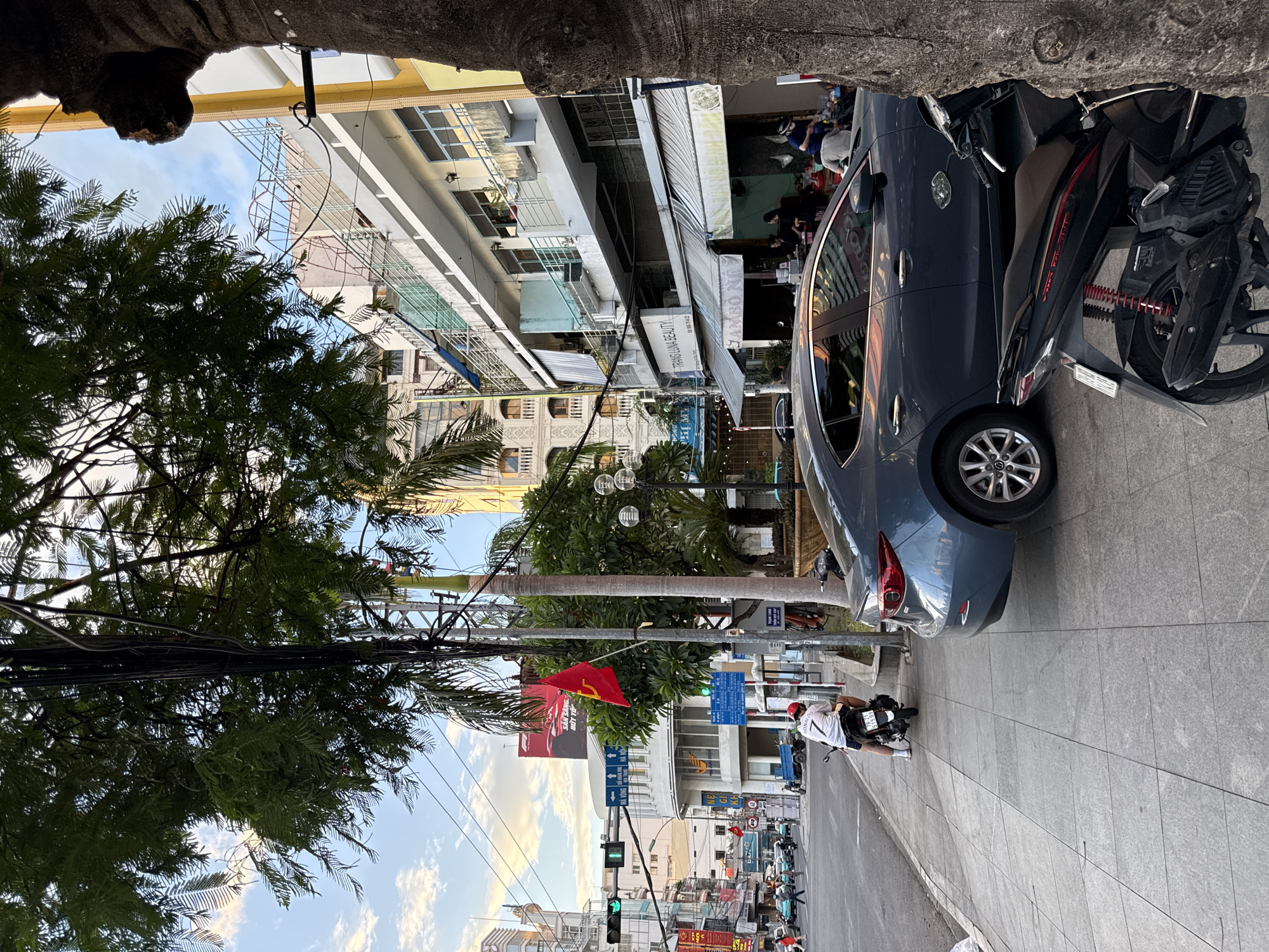 The image depicts a street scene in Nha Trang, Vietnam, likely in a commercial or mixed-use area. In the foreground, a dark-colored sedan is parked on the right side of the street, partially obscured by a large tree trunk. A black motorcycle is parked behind the sedan. To the left, a sidewalk paved with grey tiles runs alongside the street. A person wearing a white shirt with a number "7" and a red helmet is sitting on a scooter facing away from the camera, parked on the sidewalk.
The street itself is busy with traffic. Several cars and motorcycles are visible in the distance, and traffic lights are illuminated green. On the left side of the street, buildings with multiple stories line the road. One building prominently features a red flag with a yellow hammer and sickle emblem, suggesting a connection to communism. Signs in Vietnamese are visible on the buildings. Further down the street, more buildings with balconies and awnings are apparent. The sky is partly cloudy with the sun low in the sky, casting a warm light on the scene. Trees, including palm trees, are interspersed throughout the urban landscape. The overall impression is of a bustling Vietnamese city street during the late afternoon or early evening.
