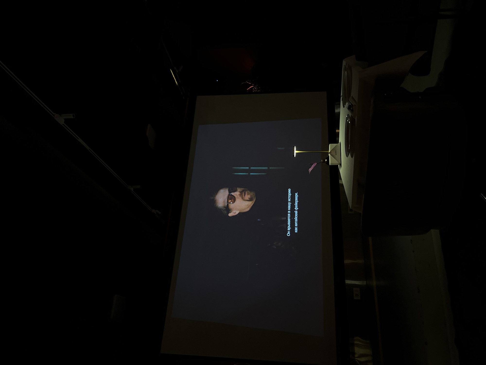 A projection screen in a darkened room displays a video of a man wearing glasses, with Russian subtitles beneath him. The environment is dimly lit, with hints of lighting fixtures and equipment visible in the periphery. The location is possibly in Yerevan, Armenia, as it is requested in the prompt.