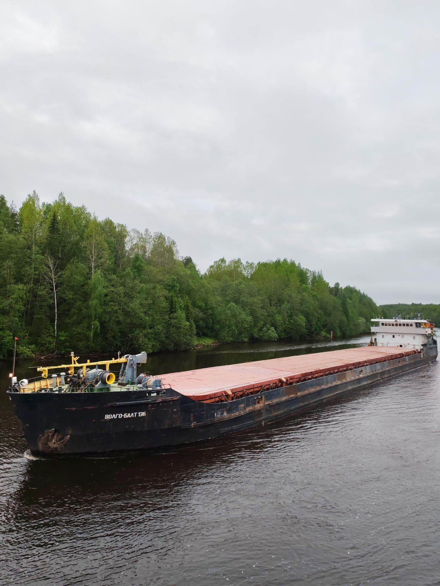 A large barge is moving along a river. The barge is black with a flat, reddish-brown deck. The barge's bow has yellow and gray equipment. The name "ВОЛГО-БАЛТ 136" is written on the side of the barge. The stern of the barge has a white, multi-story structure. The river is dark and reflective. Green trees line the riverbanks. The sky is overcast and gray. The scene suggests a location in Russia, given the Cyrillic text on the barge. The presence of the Volga-Balt waterway system suggests Depo, Russia.