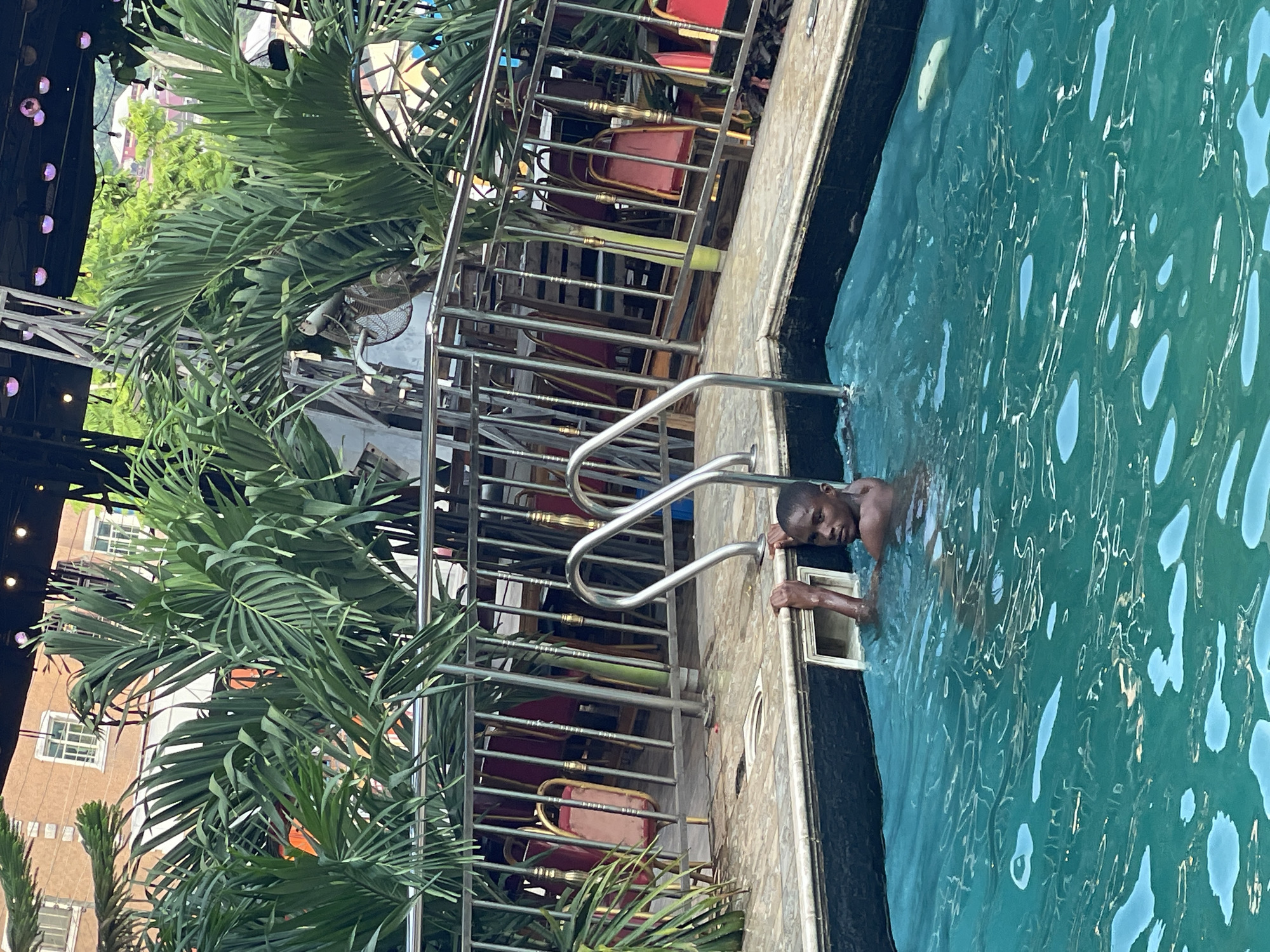 A young Black male is in a swimming pool, partially submerged, with his hands on the pool's edge. He is looking towards the camera. The pool is filled with bright turquoise water and has a white tiled edge. Behind the pool, there is a metal railing and several red chairs are visible. Palm trees with green fronds are abundant around the pool area, suggesting a tropical or subtropical environment. The background reveals a building with windows and a glimpse of greenery, indicating an outdoor setting within an urban or semi-urban location. The overall impression is a recreational scene, likely at a hotel or private residence, in Nkpor, Nigeria.