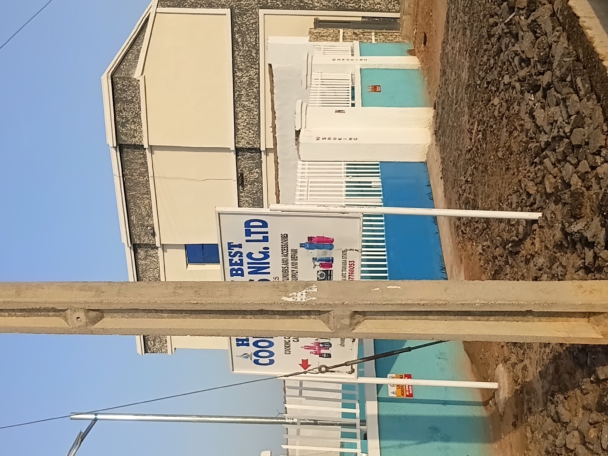 In Jalingo, Nigeria, a concrete utility pole stands in the foreground of a building with a light-colored facade and a speckled band near the roofline. To the left of the pole, a white sign displays the text "HO BEST COOKING NIG. LTD" in blue and red lettering, along with images of cooking gas cylinders and accessories. A red arrow points to the right. Behind the sign, a white fence encloses the property. The building has a blue door or window visible. To the right of the building, a white wall with vertical blue panels and a white fence suggests an entrance or gate. The ground in front of the building is covered with rough, dark-colored debris, likely rubble or asphalt. The sky is clear and blue.