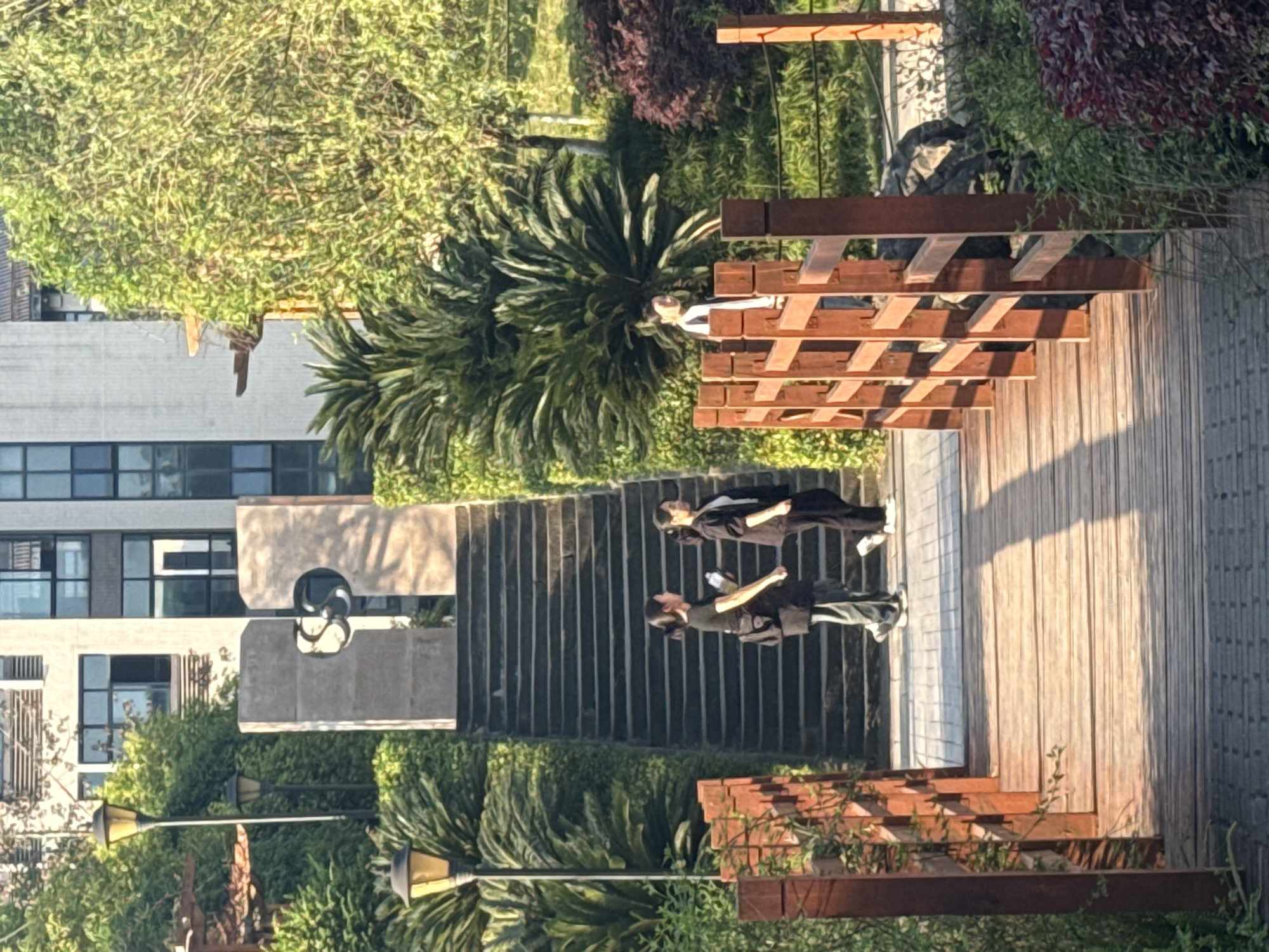 The image depicts an outdoor scene in Jiangjin, China, featuring a blend of architectural and natural elements. A building with multiple windows is visible on the left, partially obscured by trees and greenery. In the foreground, there are two individuals walking on a set of stairs adjacent to a wooden pathway. Another person is located on a wooden bridge structure. Vegetation, including trees and bushes, surrounds the pathway and building. The scene is illuminated by sunlight, casting shadows and highlighting the textures of the materials and foliage. Streetlights are present alongside the pathway.