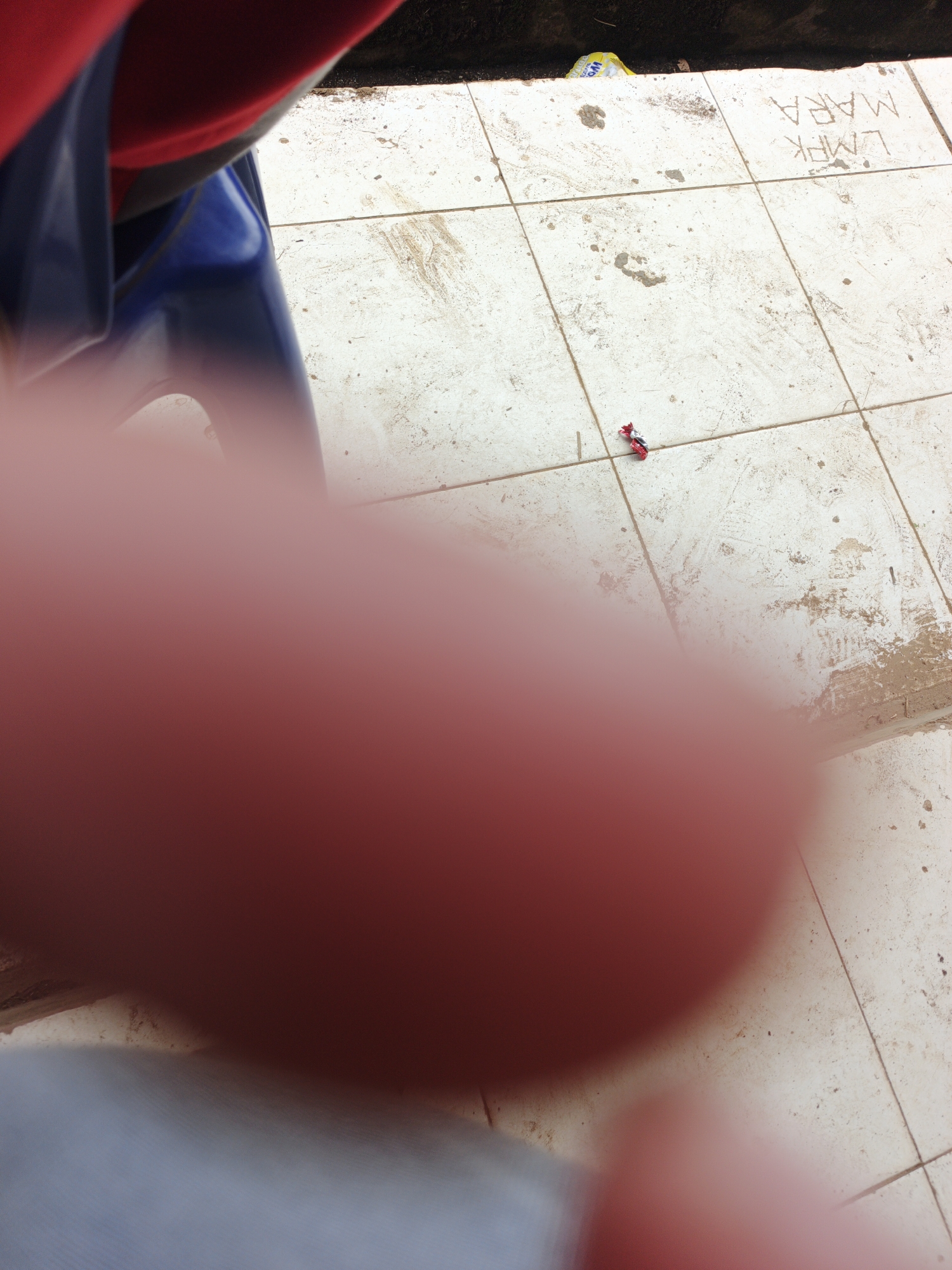 A close-up shot shows a small, red and silver toy motorcycle on a light-colored tiled floor. The floor is dirty with smudges and some scattered debris, including a yellow plastic wrapper in the background near a dark edge. To the left, part of a person's red and blue clothing is visible, as is a blurry, pinkish-red area that appears to be a thumb or finger in the foreground, obscuring a portion of the scene. In the background, faintly etched into the tiles, are the words "LMPK MARA." The setting appears to be an outdoor or semi-outdoor area, possibly a courtyard or walkway, with tiles laid out in a grid pattern. The overall impression is a casual snapshot, possibly taken by someone in the process of taking a photo of something else.