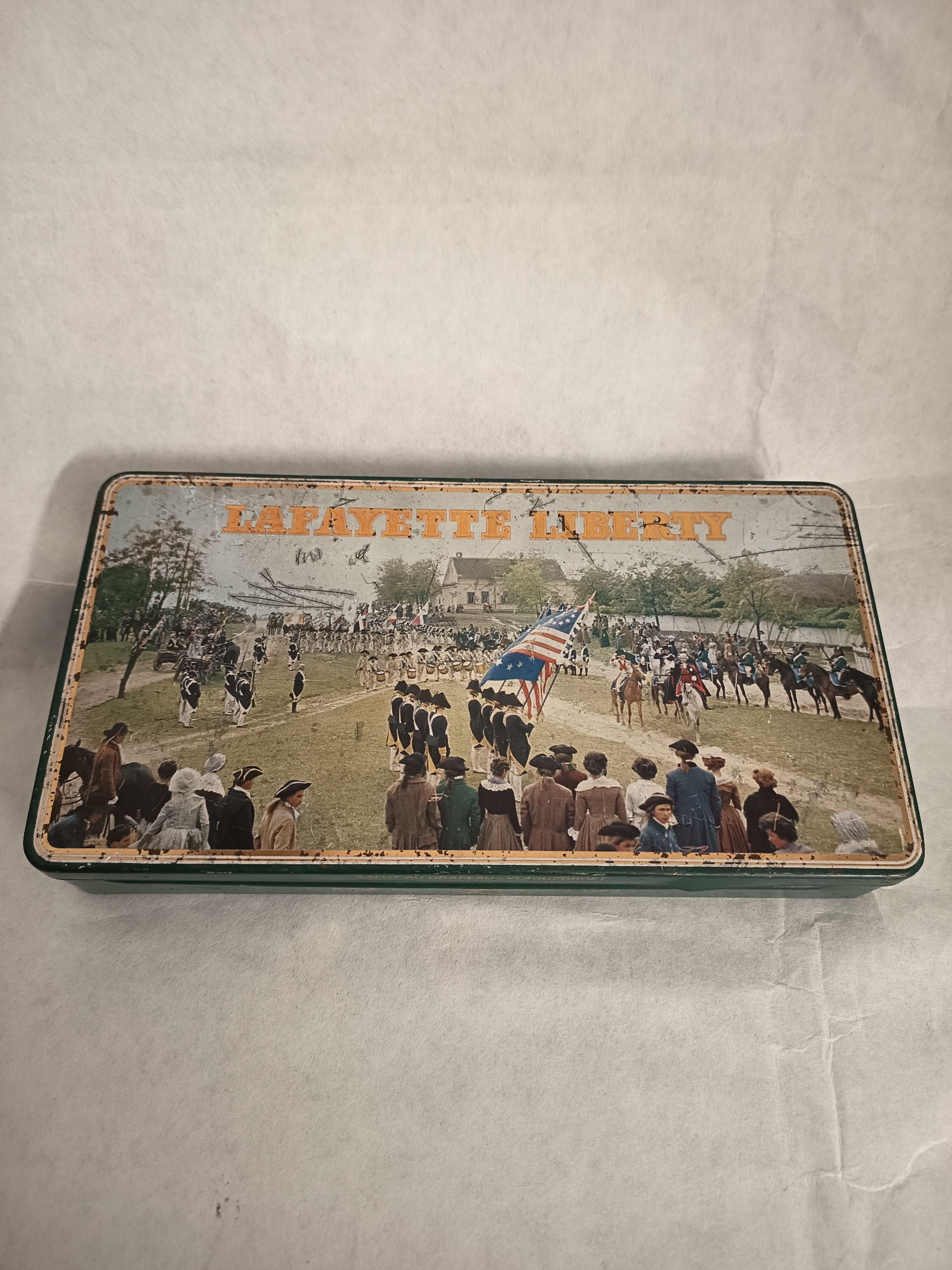 The image depicts a rectangular metal tin with a depiction of a historical scene printed on its top. The scene appears to be a parade or procession of soldiers and civilians from the late 18th or early 19th century. The people are wearing period clothing, and a large American flag is prominently displayed. A building is visible in the background.
The tin has orange lettering that reads "LAFAYETTE LIBERTY" across the top of the image. The border of the tin is dark green, and the overall appearance suggests the tin is old and has some wear.
The tin is set against a plain white or off-white background.
There is no evidence of Wangen bei Olten, Switzerland, in the image.