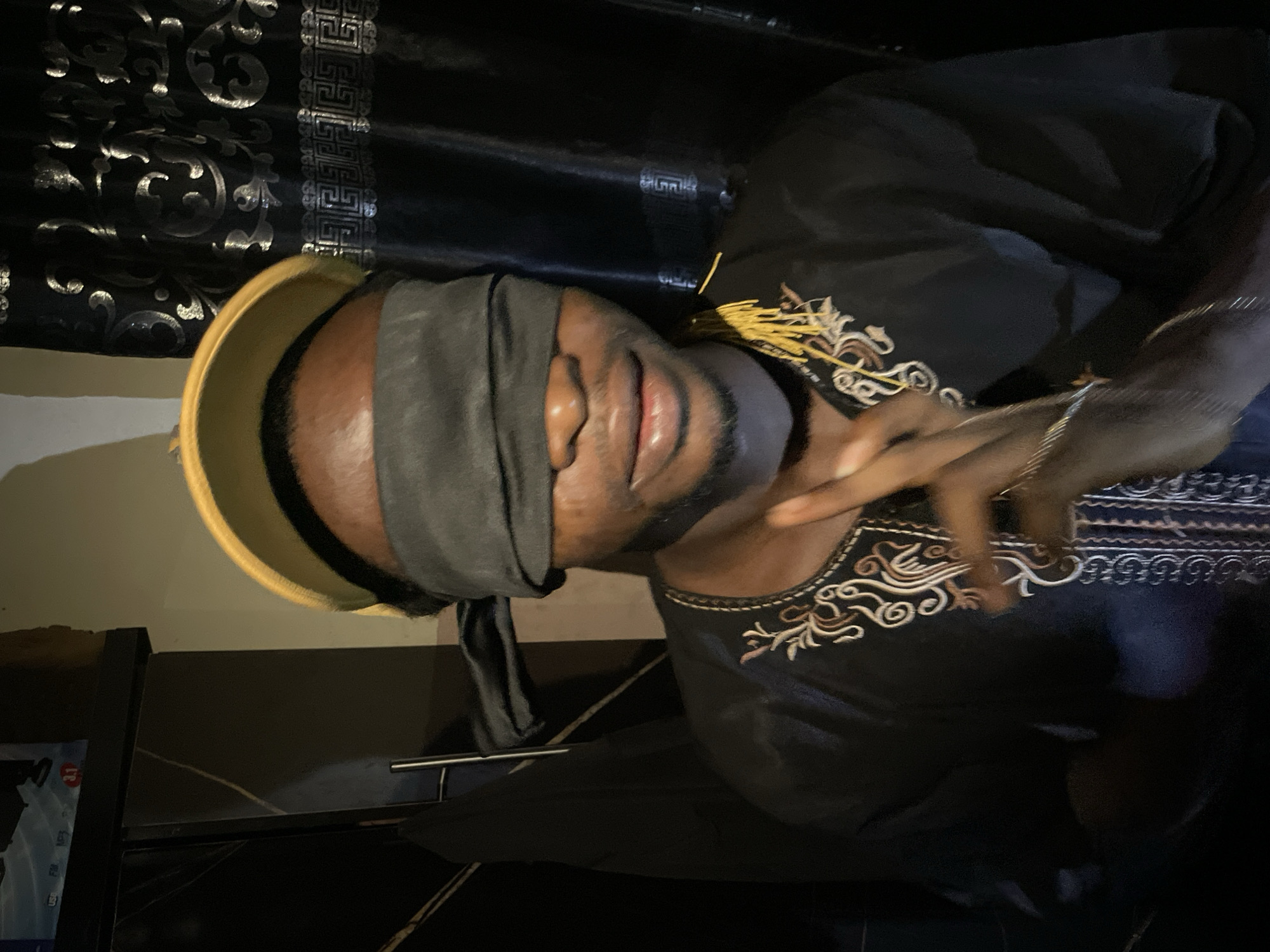 A close-up, dimly lit image shows a dark-skinned man wearing a dark-colored embroidered top and a grey blindfold over his eyes. A gold-colored rimmed object sits atop his head, possibly a bowl or hat. He is gesturing with his right hand. The background is dark with patterned curtains visible in the top left corner. The location, Lokoja, Nigeria, cannot be confirmed based on the visual content of the image. The presence of a black shelf or table and a glimpse of a white and blue poster are also observed in the lower left area of the image.