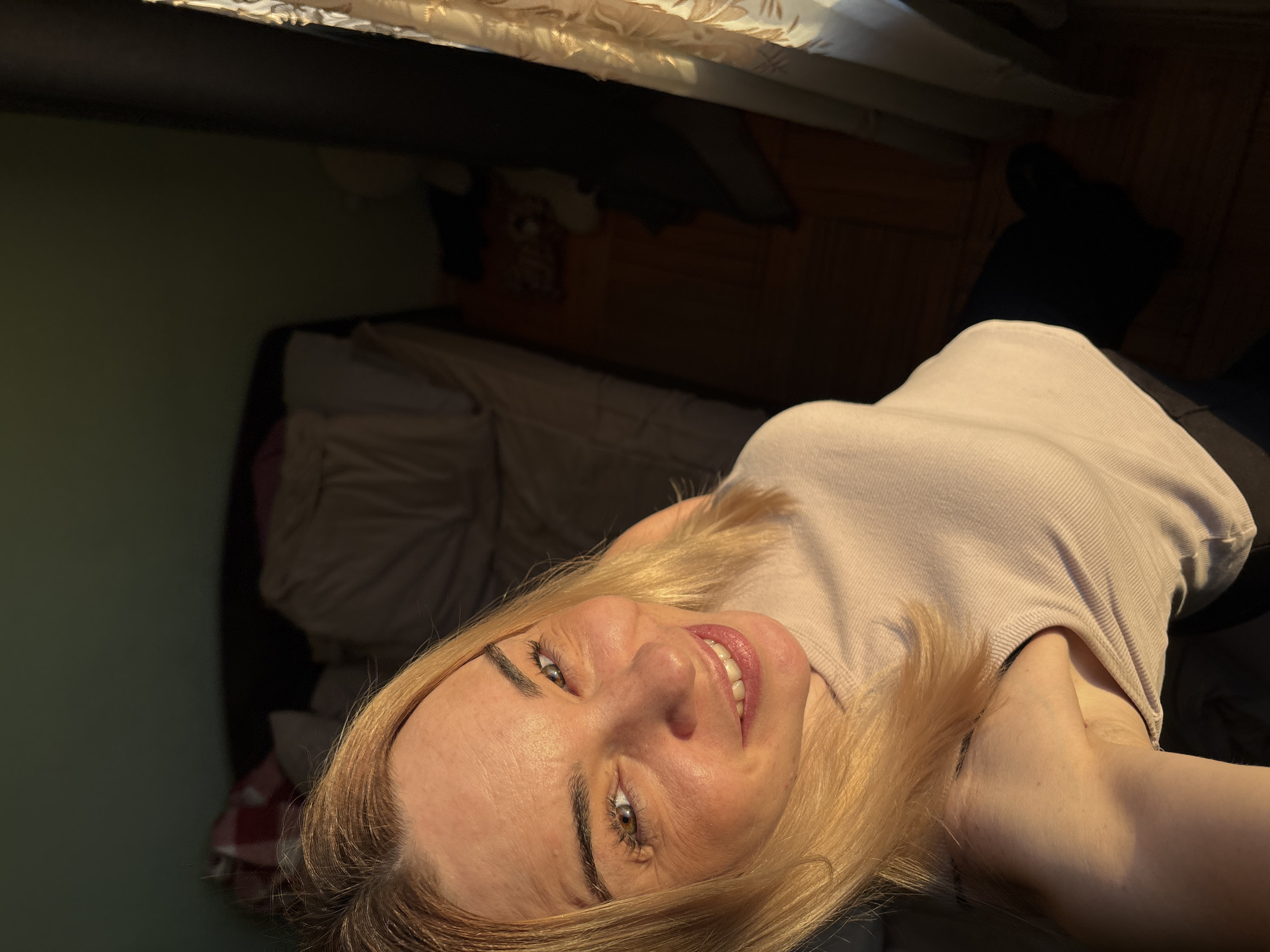 A blonde woman with fair skin and light brown/hazel eyes is shown in a selfie-style portrait, looking directly at the camera with a gentle smile. The image is rotated counter-clockwise, making her appear to be looking upwards. She is wearing a light-colored, possibly beige or pale pink, ribbed sleeveless top.
Strong, warm natural light illuminates her face and blonde hair from the upper right, creating highlights and shadows that define her features. Her hair is cut short, appearing layered around her face. She has well-defined eyebrows, and her lips are slightly parted, revealing her teeth.
The setting is an indoor room, likely a bedroom or living space. In the background, a light green wall is visible on the left, transitioning to a darker, more cluttered area with a dark wooden floor visible on the right. A patterned curtain or fabric is present at the top of the frame, possibly indicating a window or part of a bed. Various indistinct, dark items, potentially clothing or furniture, are visible in the background, mostly out of focus. The overall mood is pleasant and casual.
No visible text is present in the image.