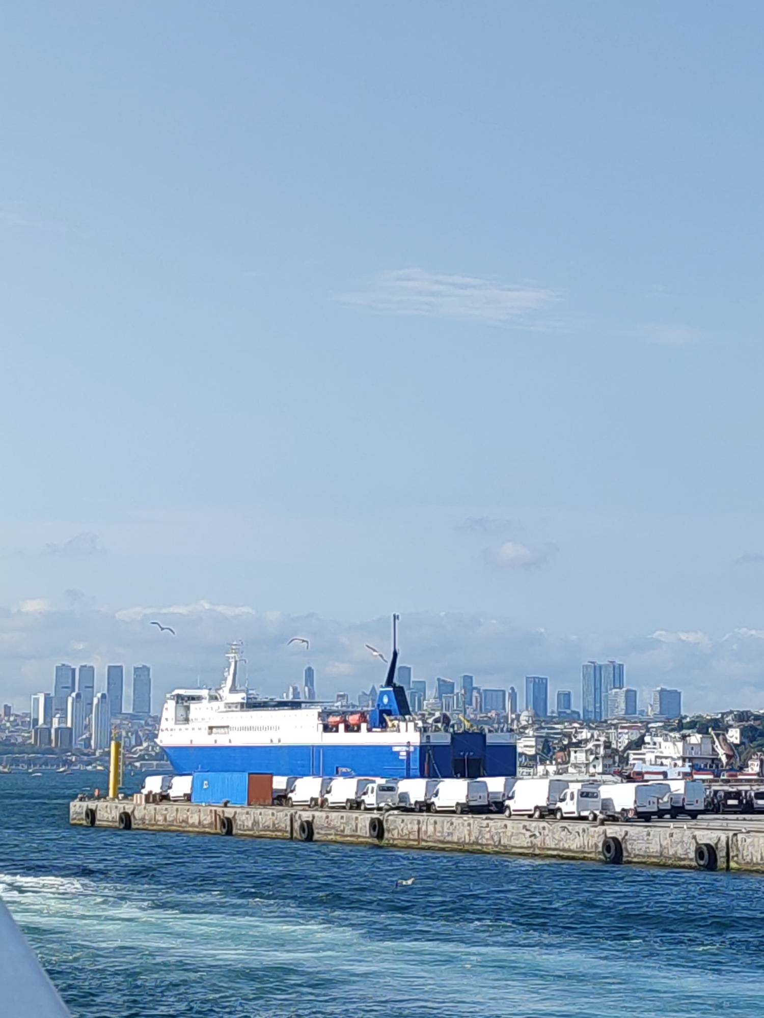 The image shows a ferry docked at a pier in Istanbul, Turkey, with a cityscape in the background. The ferry is large and predominantly blue and white, with several vehicles, including white vans and a blue container, parked on its deck. The pier is made of concrete and lined with tires. Several seagulls are visible flying in the sky. The water is a deep blue with white foam indicating movement. The cityscape features numerous tall buildings, suggesting a densely populated urban area. The sky is clear blue with a few scattered clouds. The scene appears to be a sunny day.
