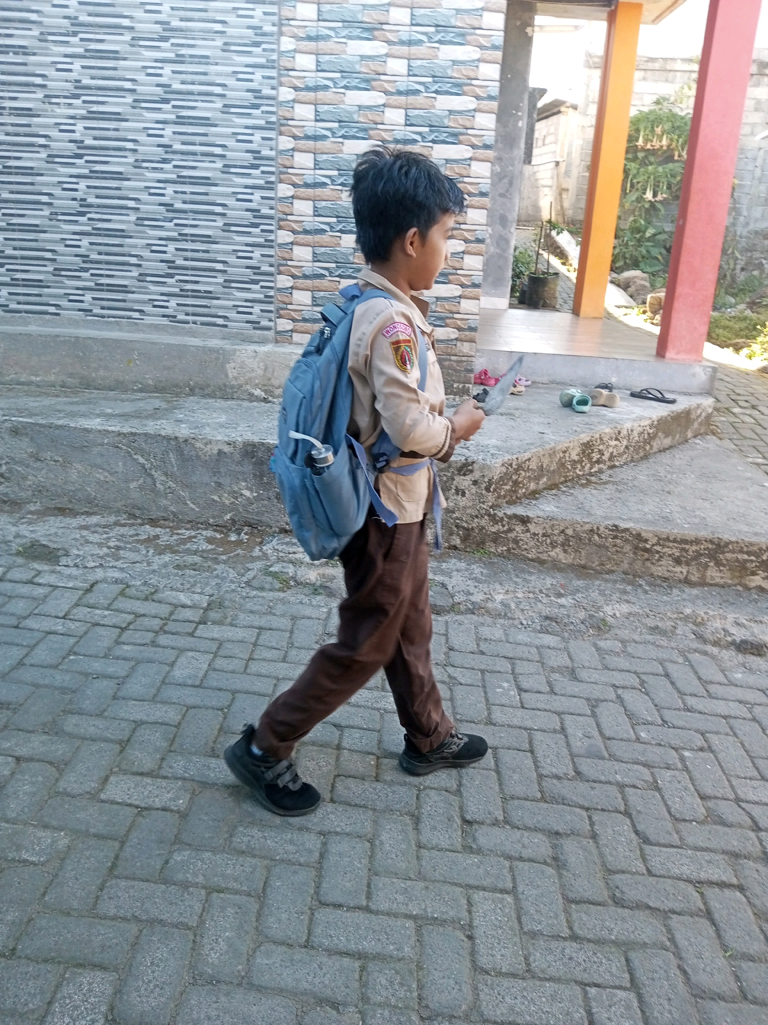 In Andongsili, Indonesia, a young boy in a scout uniform walks across a brick pathway. He carries a blue backpack with a water bottle in the side pocket. He is holding a knife in his right hand and appears to be looking at it. The background includes a tiled wall, a concrete staircase, and several pairs of footwear. A building with orange and red pillars is visible in the distance.