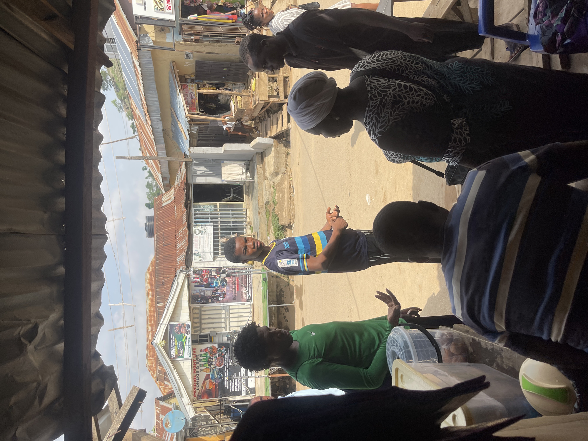 Here's a concise description of the image:
The image shows a street scene in Abuja, Nigeria, likely captured from inside a covered stall or shop. Several people are present. A woman wearing a headscarf and patterned dress is among them. A young man in a sports jersey stands on the street. Another person wears a green shirt. Buildings line the street, with metal roofs and walls displaying signs and advertisements. One sign advertises "IMPERIAL NIGHT LIVE LONUGE & BAR". The general environment appears to be a busy commercial area.