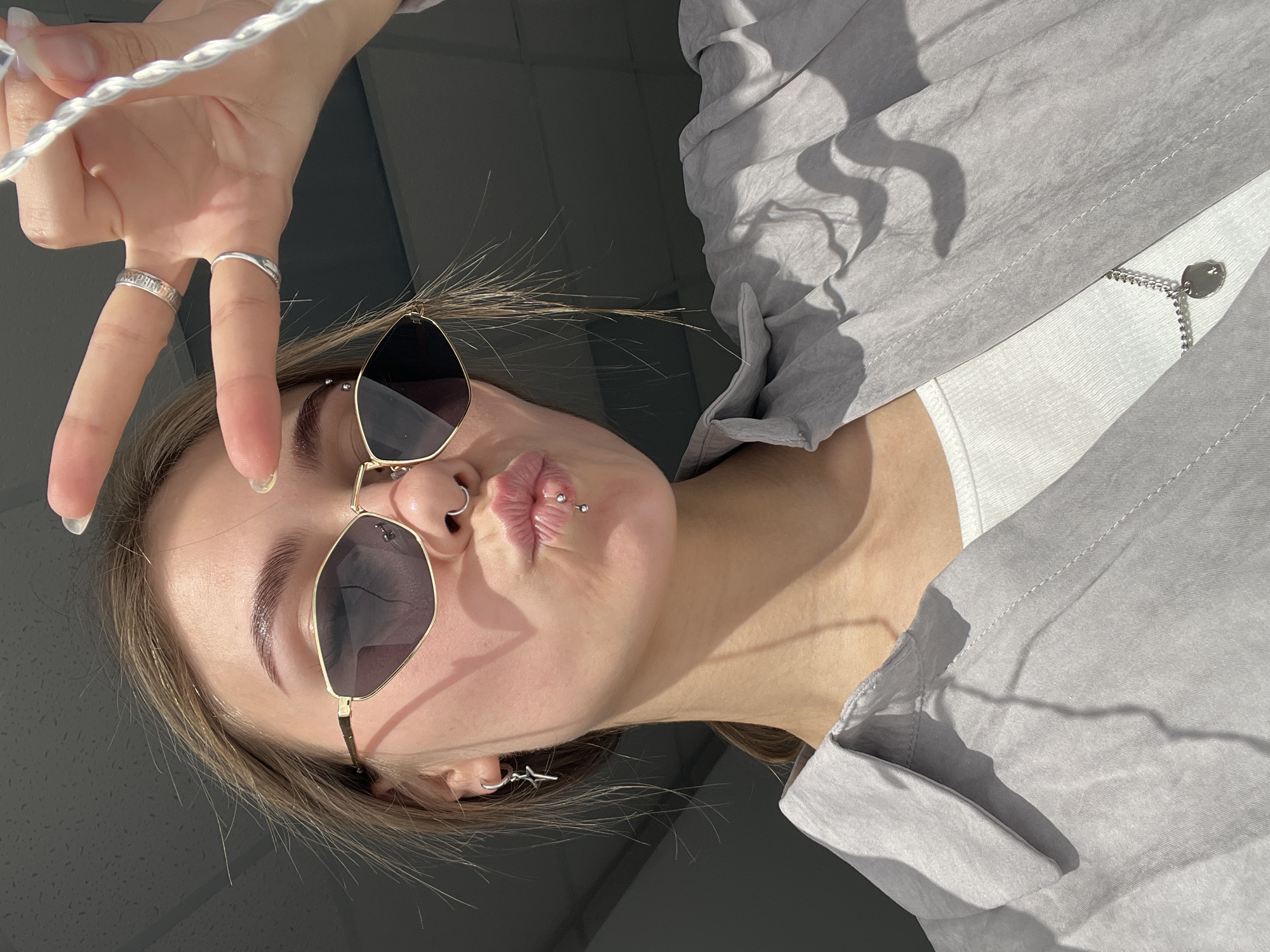 A woman takes a selfie. She is wearing sunglasses and has lip and nose piercings. She is making a peace sign with her hand. She is wearing a grey shirt. The location cannot be determined from the image.
