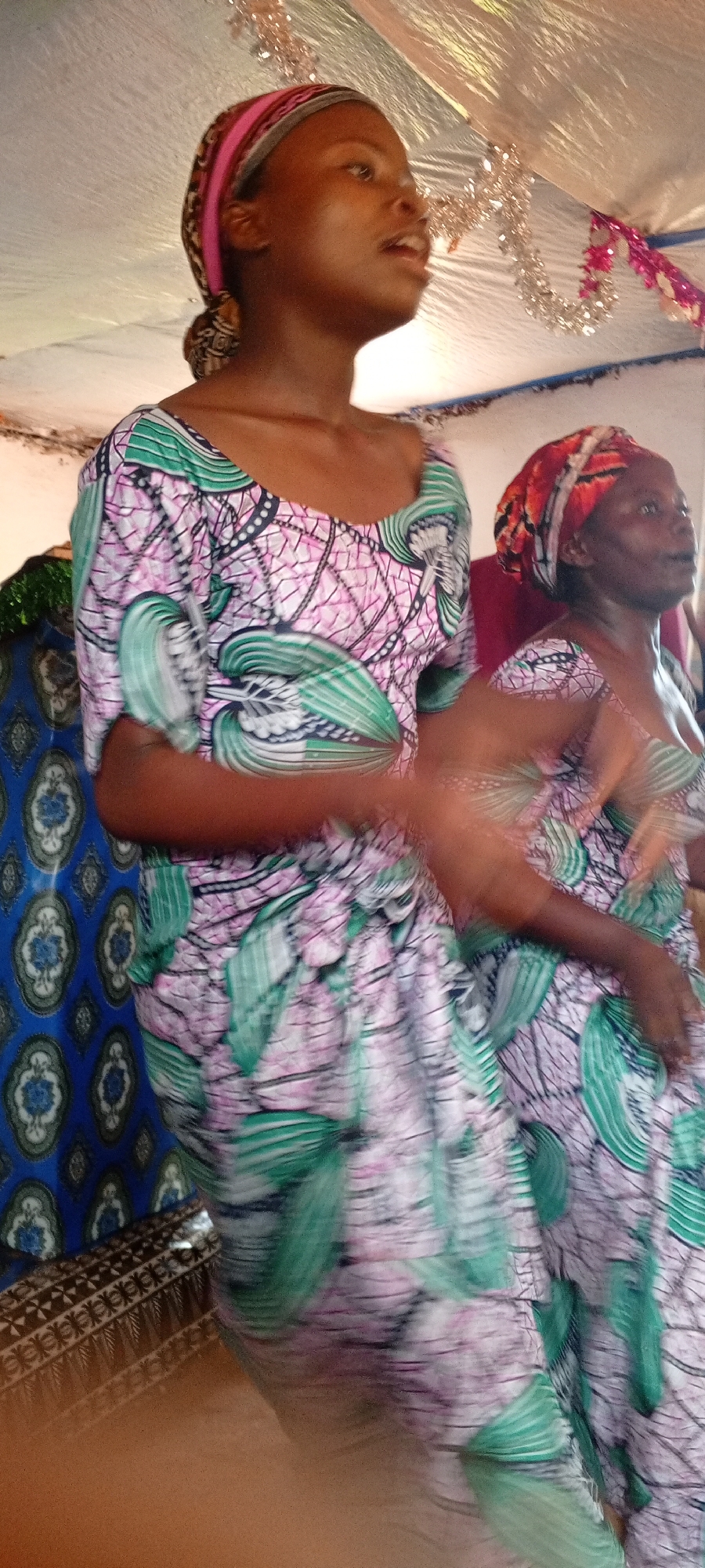 In a blurred indoor shot, two women stand adjacent to each other under what appears to be a tent or draped canopy with metallic streamers. Both are wearing similar dresses with a repeating pattern of green leaf-like shapes over a pink and white base. The woman in the front is younger, wearing a matching headscarf, and is looking upwards with her mouth open, possibly singing or speaking. The woman behind her, also wearing a matching headscarf, is older and appears to be looking upwards as well. To the left, a blue patterned fabric is visible. The location is identified as Makungu, Democratic Republic of the Congo.