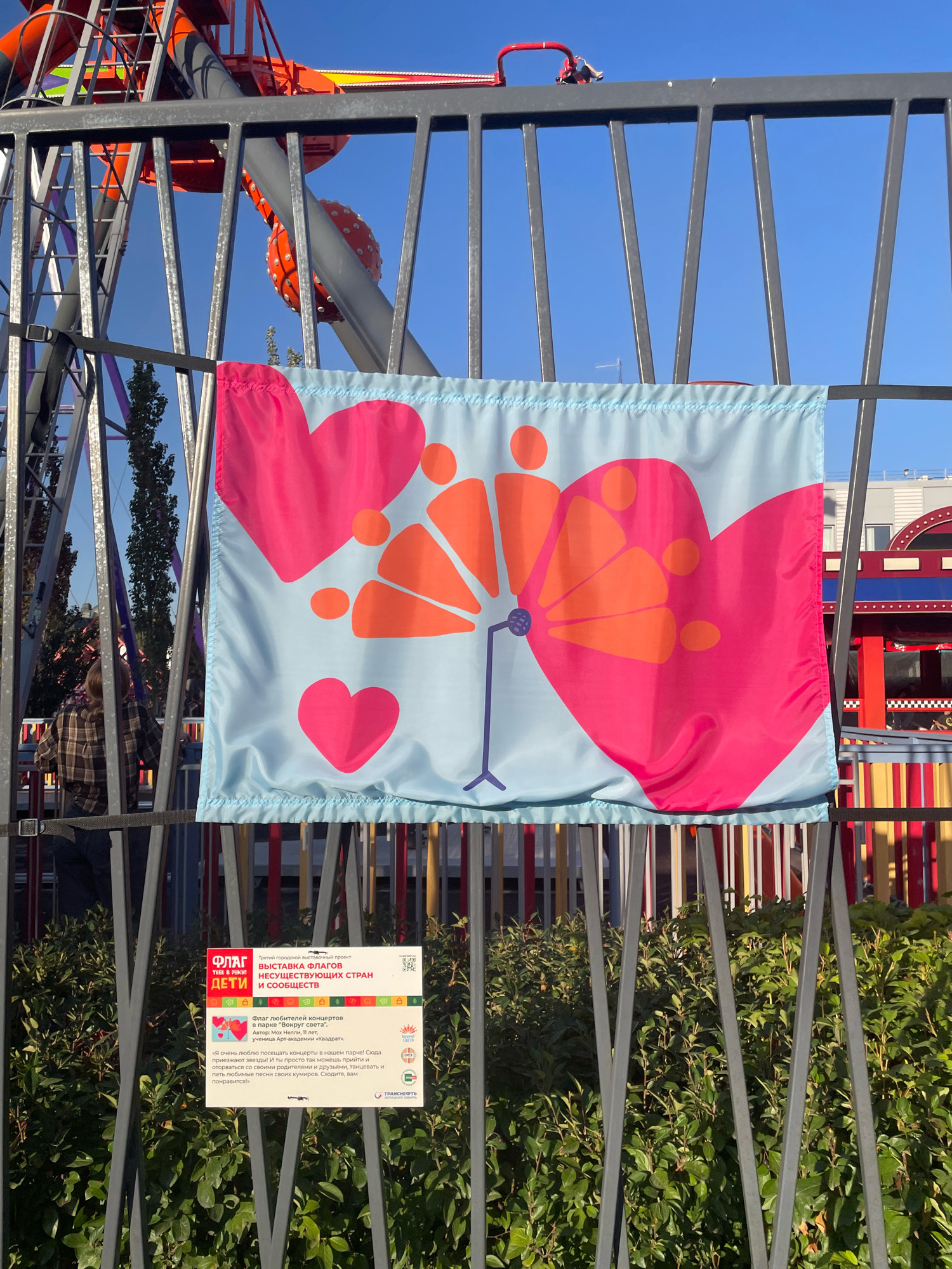 A light blue flag with pink and orange heart and flower designs hangs on a dark metal fence. To the left of the flag is a portion of a Ferris wheel or similar amusement park ride with orange and gray structures and a blue sky. To the right of the flag, beyond the fence, is a colorful attraction with red and yellow striped poles. Below the flag, attached to the fence, is a white sign with text and images. The sign has Cyrillic text and logos, and one of the logos appears to be the city of Omsk. The flag features abstract shapes of hearts and a stylized flower, with smaller orange circles and shapes. The fence has vertical metal bars and a black strap is attached to the lower left. Green bushes are visible below the fence.