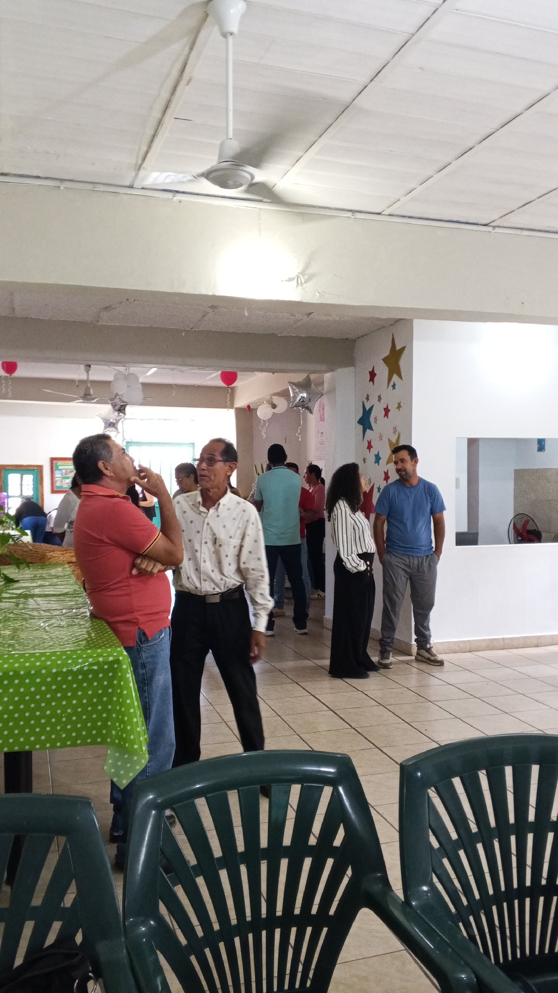 The image depicts an indoor gathering of people in what appears to be a hall or community center. Two men are standing near a table covered with a green polka-dotted tablecloth, engaged in conversation. One man is wearing a red shirt and jeans, while the other wears a white patterned shirt and black pants. Further back, a woman in a striped shirt and black pants stands near a man in a blue shirt and gray pants. Other individuals are visible in the background, near red and silver balloons and star decorations adorning the wall. The environment features tiled flooring, white walls, and a white ceiling with ceiling fans. Two green plastic chairs are in the foreground.