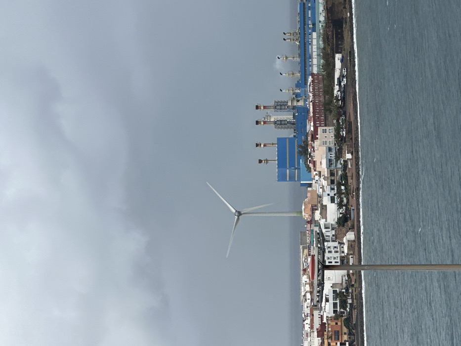 An elevated long shot captures a coastal industrial and residential landscape in Puerto del Rosario, Fuerteventura, Spain, under a heavily overcast, grey sky. In the foreground, choppy, dark blue-green ocean water occupies the lower quarter of the frame. A tall, slender grey pole with a multi-tiered platform topped by a red dome stands vertically in the left foreground, partially obscuring the view of the shore.
The midground features a dense array of residential buildings, predominantly white and pastel-colored, lining a rocky coastline. Several parked vehicles are visible on streets between the buildings. Behind and to the right of the residential area, a large industrial complex, identified as a power plant, dominates the background. It comprises extensive blue and grey structures with multiple tall smokestacks, some emitting faint plumes of white smoke or steam. To the left of the power plant, a prominent white wind turbine with three rotating blades rises above the residential structures. No individuals are discernible within the scene.