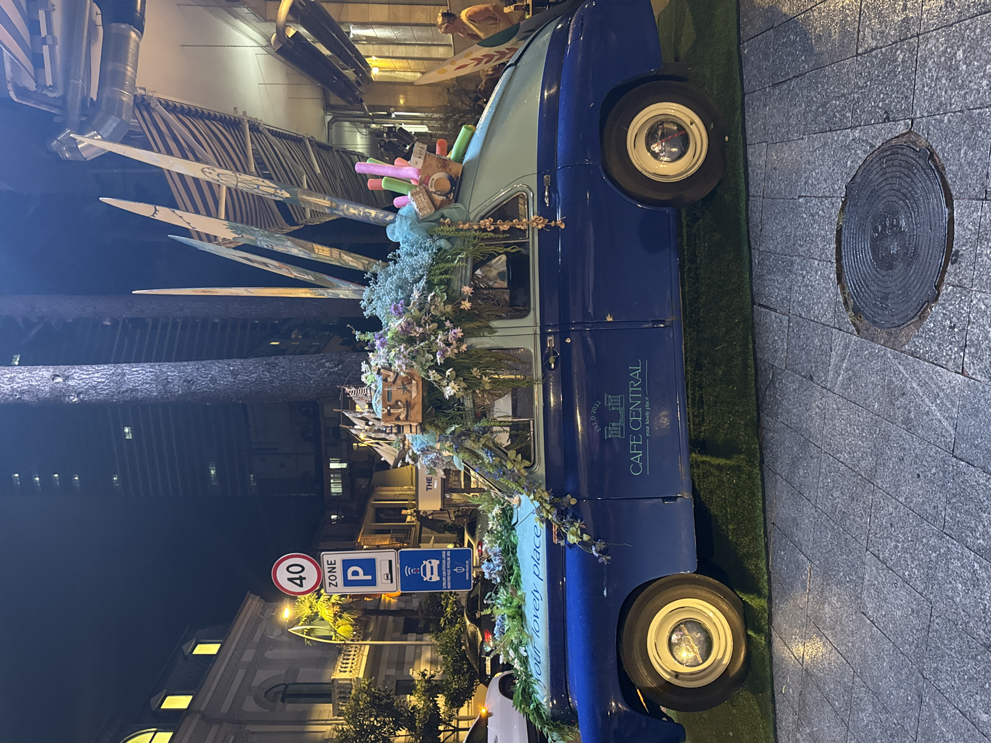 This is a night time photograph of a vintage blue car decorated with flowers and decorative items, parked on a cobblestone street in Batumi, Georgia. The car has "Cafe Central - your lovely place" written on its side. The scene appears to be an outdoor seating area for the cafe. In the background, a tall modern building with illuminated windows and an older building with arched windows can be seen. Traffic signs are visible on the left, indicating a speed limit of 40 and parking. A person is visible in the background on the right, standing near a table. The car is adorned with a large floral arrangement on the roof and a small wooden boat-like structure. Various colorful cylindrical objects are also attached to the roof. The ground is covered with artificial grass in front of the car, leading to the cobblestone pavement. A manhole cover is visible in the foreground.