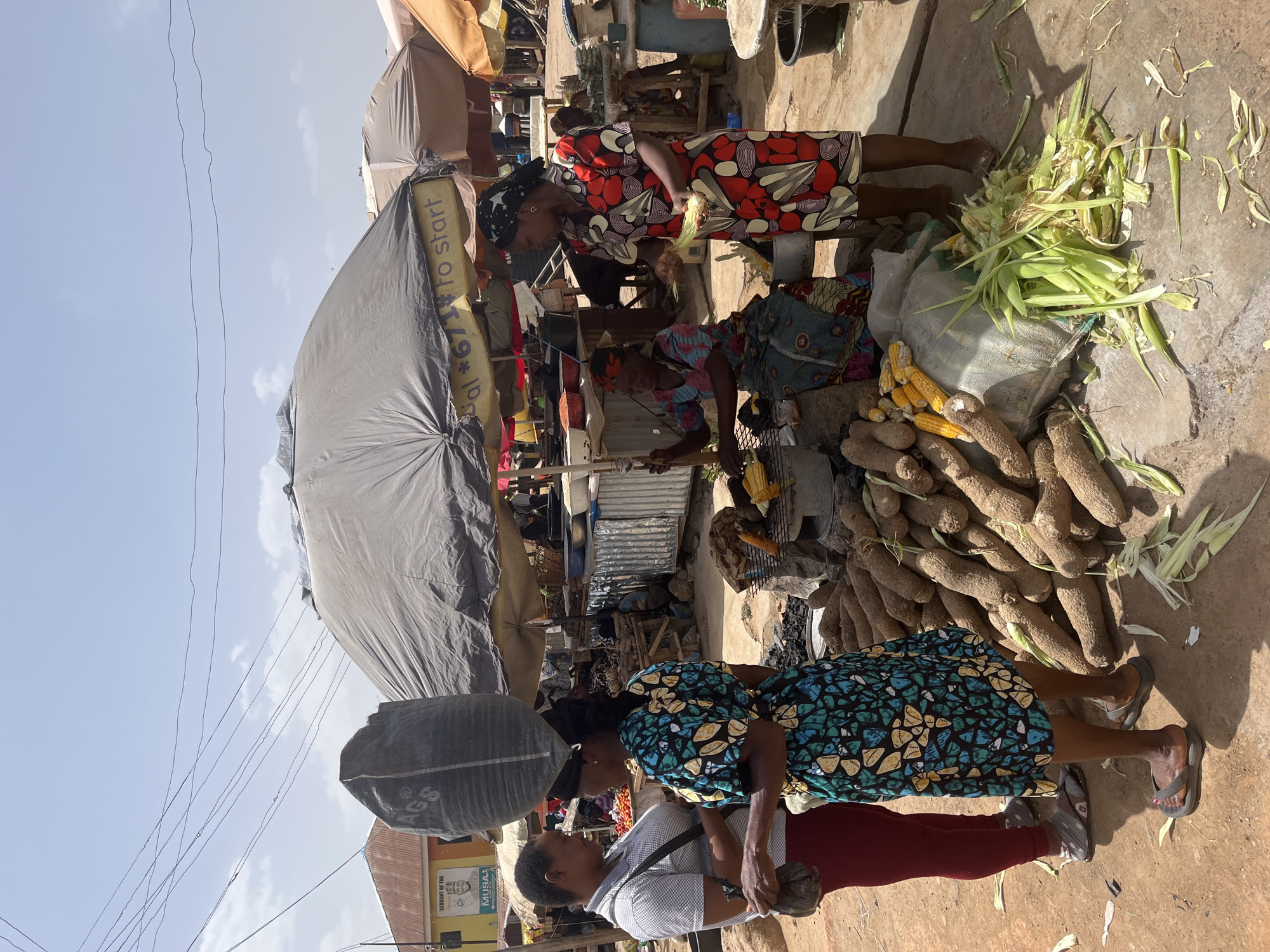 The image depicts a vibrant market scene in Gwong, Nigeria. Two women are selling agricultural produce, including yams and corn. One woman wears a colorful red and white patterned dress, while the other wears a blue and yellow patterned dress. One is husking corn. A large pile of yams and a bag of corn are on the ground. Another woman walks by with a heavy load balanced on her head, interacting with someone in a white shirt and red trousers. The market is under shade provided by a large umbrella and other coverings. In the background, there are various stalls and signs of the bustling urban environment. Electrical wires run across the clear sky above the market.