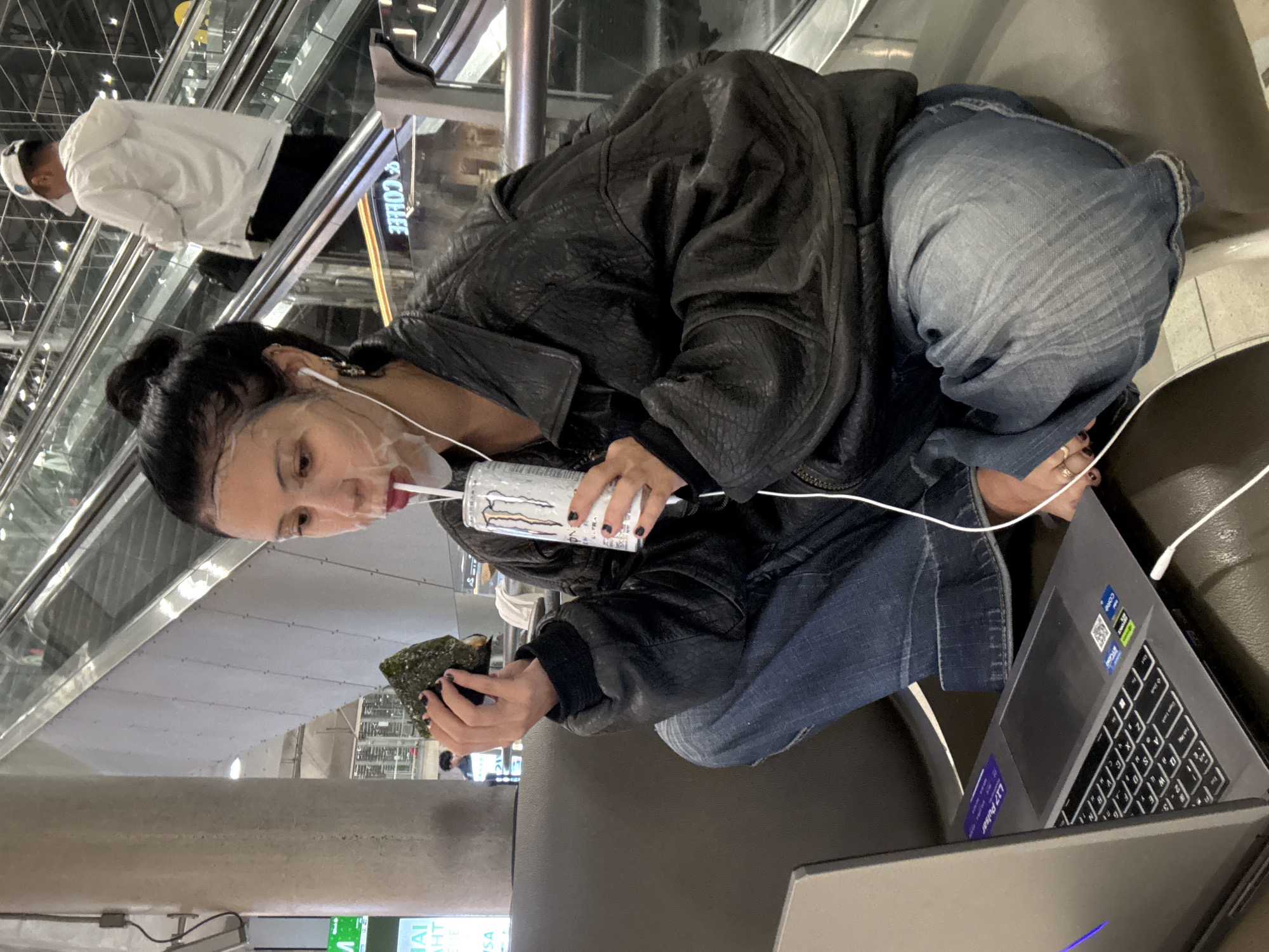 A young adult with dark hair pulled back in a bun, wearing a face mask and a black leather jacket over a black t-shirt, sits cross-legged on a brown bench in what appears to be an airport terminal. They are holding a piece of nori seaweed in their left hand and a can of Monster Energy drink in their right hand, with a straw inserted into the can. A laptop is open on the bench in front of them, and white earbuds are in their ears, with the wire trailing down. In the background, an escalator is visible with people on it, and a coffee shop sign with the word "COFFEE" in reverse is on the right side of the image. The lighting suggests an indoor environment.