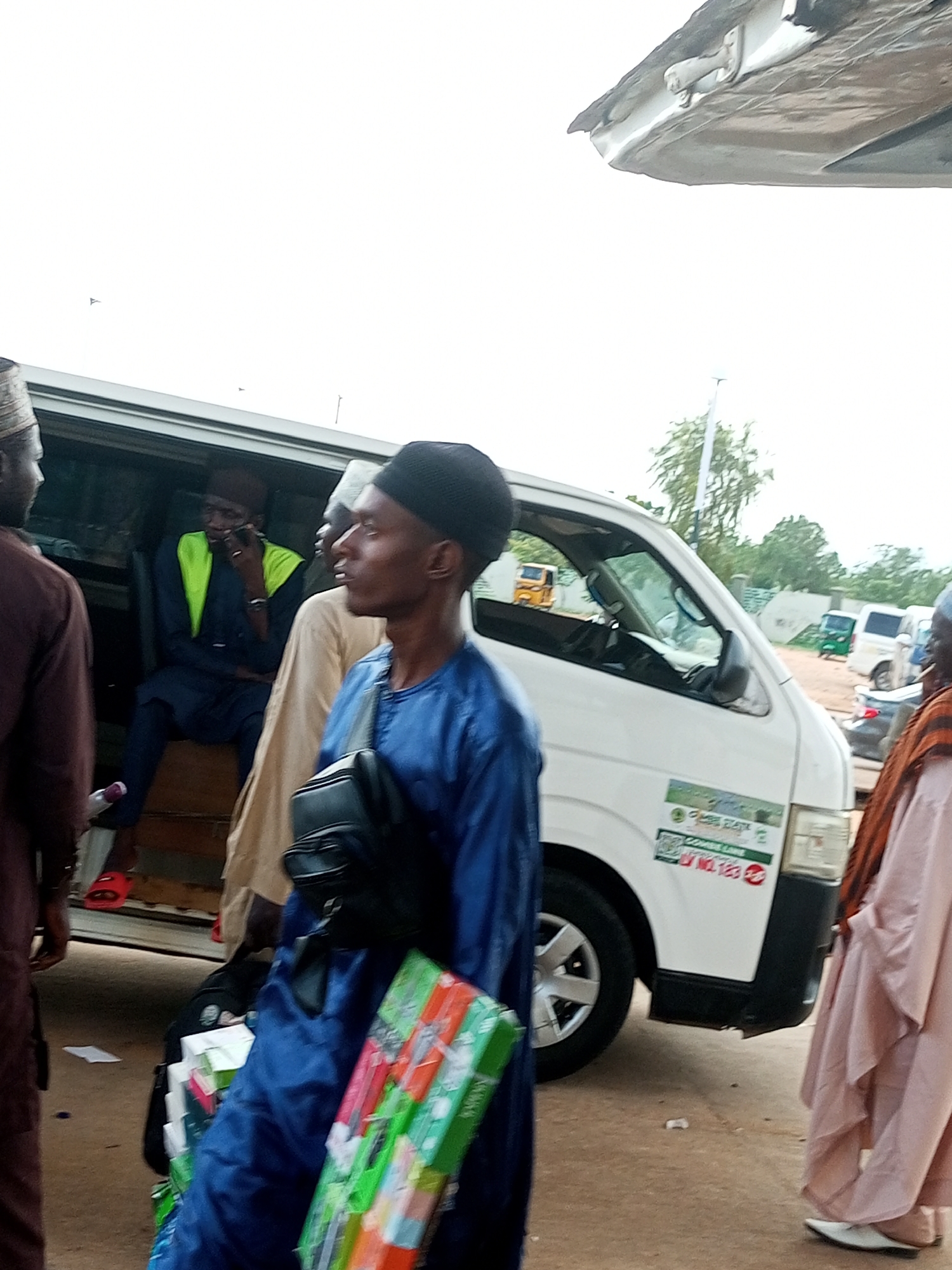 This image depicts a scene likely at a motor park in Gombe, Nigeria, possibly near the Gombe Central Market, given the presence of a commercial bus and people dressed in traditional attire. The primary subject is a white commercial bus, identified by a sticker reading "Gombe Lines" and "L/NO. 123". Several individuals are visible. In the foreground, a man in a blue outfit and a black cap is looking to the side, holding a bag with items that appear to be boxed goods with colorful packaging. To his left, another person in a brown garment is partially visible. Inside the bus, a man in a blue outfit with a bright green vest is seated and engaged in a phone conversation. Another person in a light-colored garment is standing next to him. To the right of the bus, a person in a light pink robe and white shoes is also visible. The ground is dusty, suggesting an outdoor environment common in Nigerian cities. The overall atmosphere appears to be one of transit and daily commerce.