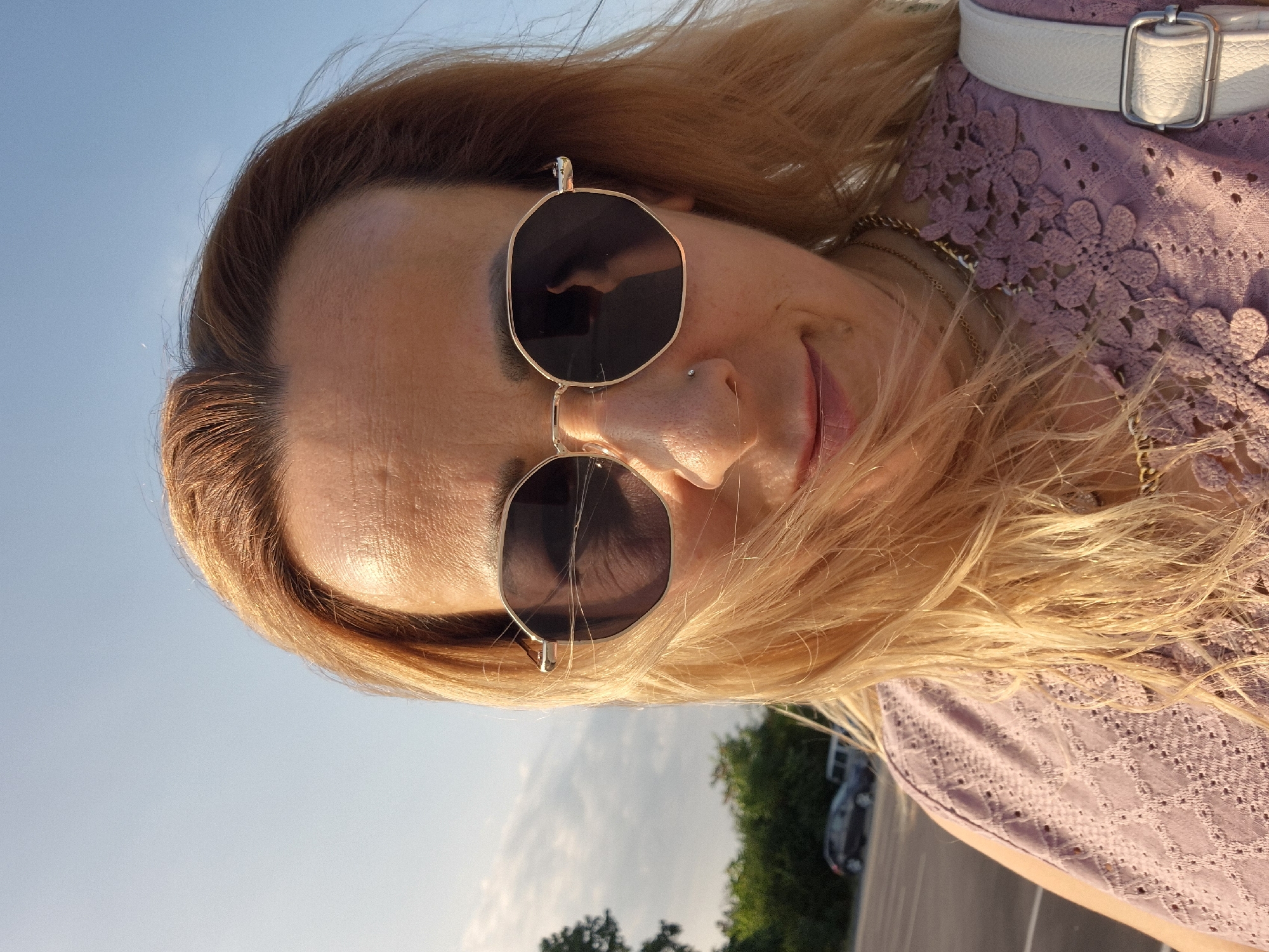 A woman with blonde hair and sunglasses is taking a selfie outdoors. She is wearing a purple lace top and a white purse strap is visible. In the background, there is a parking lot with cars and trees under a blue sky. The sun is shining. The location is Unknown, Unknown.