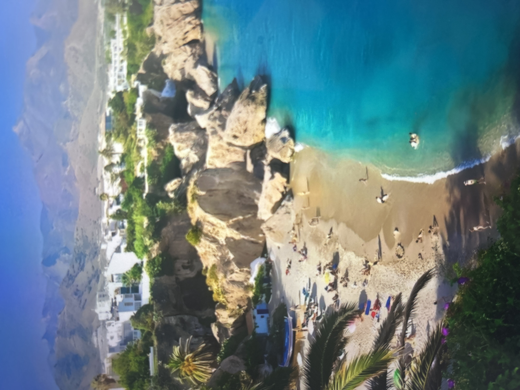 An aerial or high-angle shot captures a vibrant coastal scene under bright daylight. The image is rotated, presenting a beach at the bottom, a town on the hillside to the right, and distant mountains to the far right.
The central subject is a sandy beach, bustling with numerous people. Some individuals are visible sunbathing on towels or lying on the sand, while others are walking near the water's edge. A few people are swimming or wading in the clear, turquoise ocean close to the shore. Beach umbrellas in various colors, including blue and red, are scattered across the sand, providing shade for beachgoers. A small blue boat or paddleboard rests on the sand near the cliffs.
To the right, prominent rocky cliffs and outcrops frame the beach, separating it from a cluster of white buildings with flat roofs, characteristic of Mediterranean architecture. These structures appear to be part of a town or resort, nestled amidst lush green vegetation and palm trees. Further in the background, hazy, arid mountains rise under a clear blue sky.
The ocean displays a beautiful gradient of colors, from light turquoise near the sandy shore where gentle waves break, to a deeper blue further out. One or two individuals can be seen swimming or floating in the deeper water. The strong sunlight casts distinct shadows, suggesting a midday or early afternoon setting. The overall scene conveys a lively, relaxing holiday atmosphere. No visible text is present in the image.