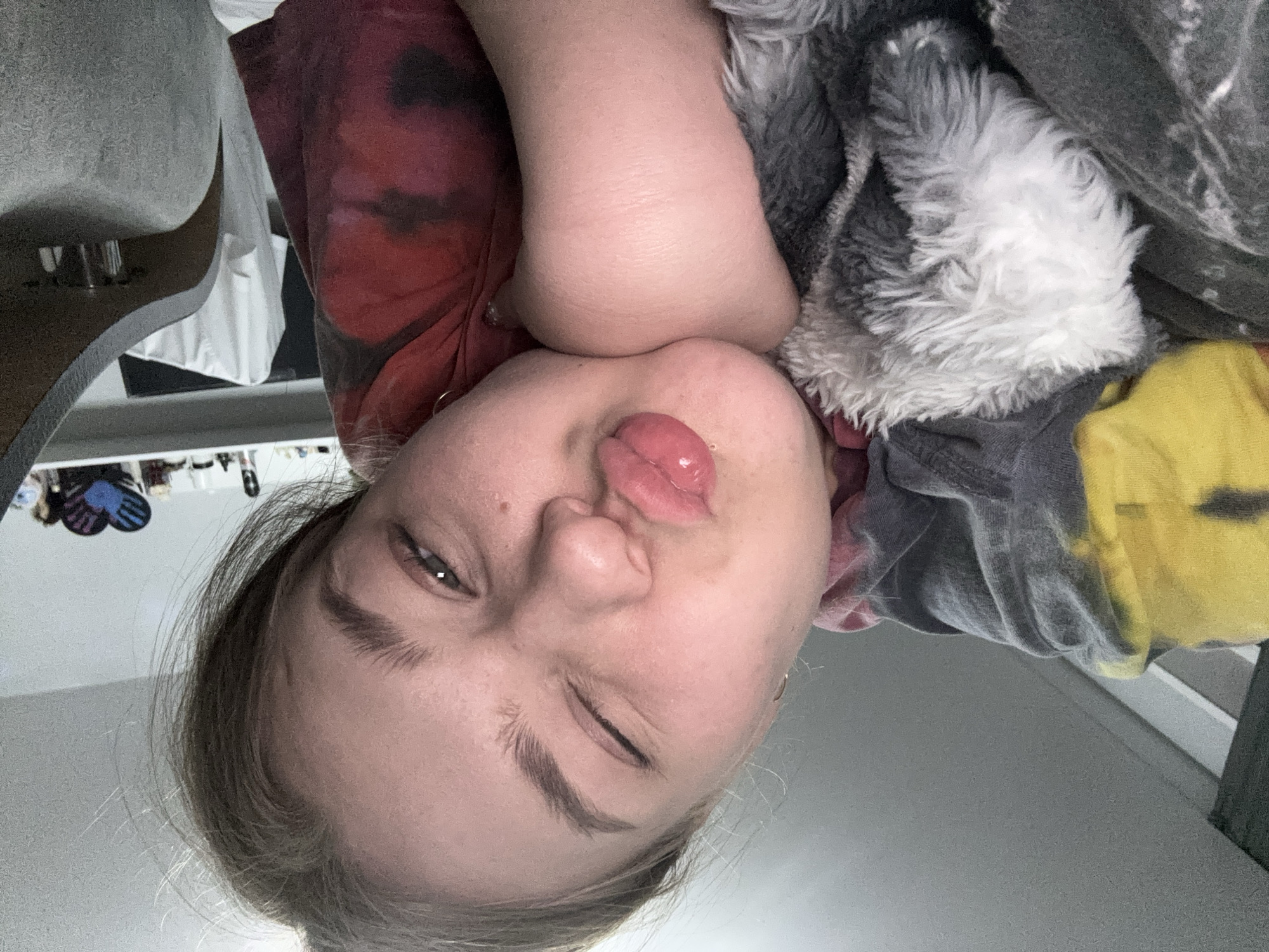 A selfie of a young person is captured at a tilted, upside-down angle. The person is fair-skinned and has light-colored hair, which is partially visible in the bottom left corner of the frame. Their left eye is closed in a wink, and they are puckering their lips in a kissing gesture. The person's head rests on their right arm, which is draped over a grey and white fuzzy blanket. A red and black tie-dye shirt is visible on the left side of the frame. The background includes a guitar, a white dresser, and a grey wall. A yellow shirt is visible on the right side of the frame. The location cannot be confirmed as Chaikovsky, Russia, based on the provided image.