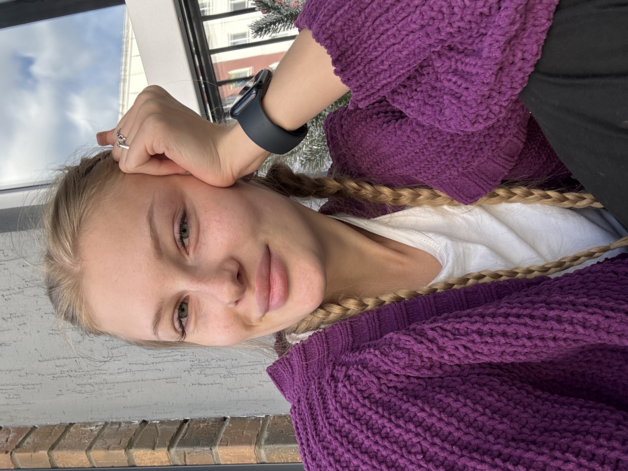 A young woman with blonde hair in pigtails, freckles, and green eyes is captured in a close-up, outdoor selfie. She is wearing a thick, purple knitted cardigan over a white t-shirt and resting her chin on her hand, adorned with a ring. A dark smartwatch is visible on her wrist. The background features a textured grey wall, a brick column, and a large window with a cloudy sky and building reflections. The scene appears to be in Perm, Russia.