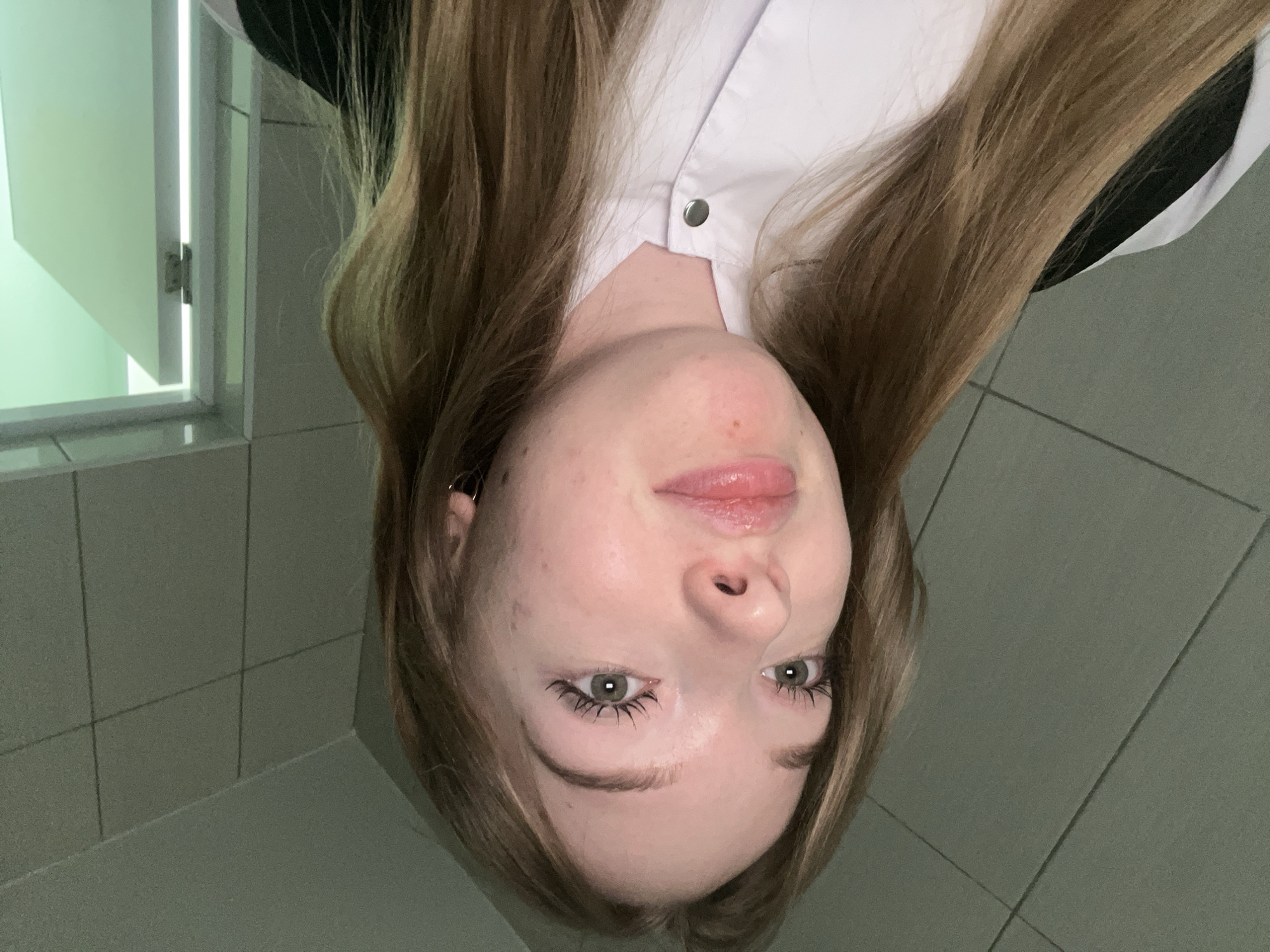 A close-up, upside-down photograph shows a fair-skinned young woman with long, light-brown hair, wearing a white shirt. Her facial features are centered in the frame; she has light-colored eyes with dark lashes. The background appears to be a tiled wall, suggesting a bathroom or similar utilitarian space. A partially open cabinet with a glossy green surface is visible in the upper left corner. The lighting appears to be fluorescent or artificial. It is not possible to determine the location in the city of Chaikovsky, Russia, from this single image.
