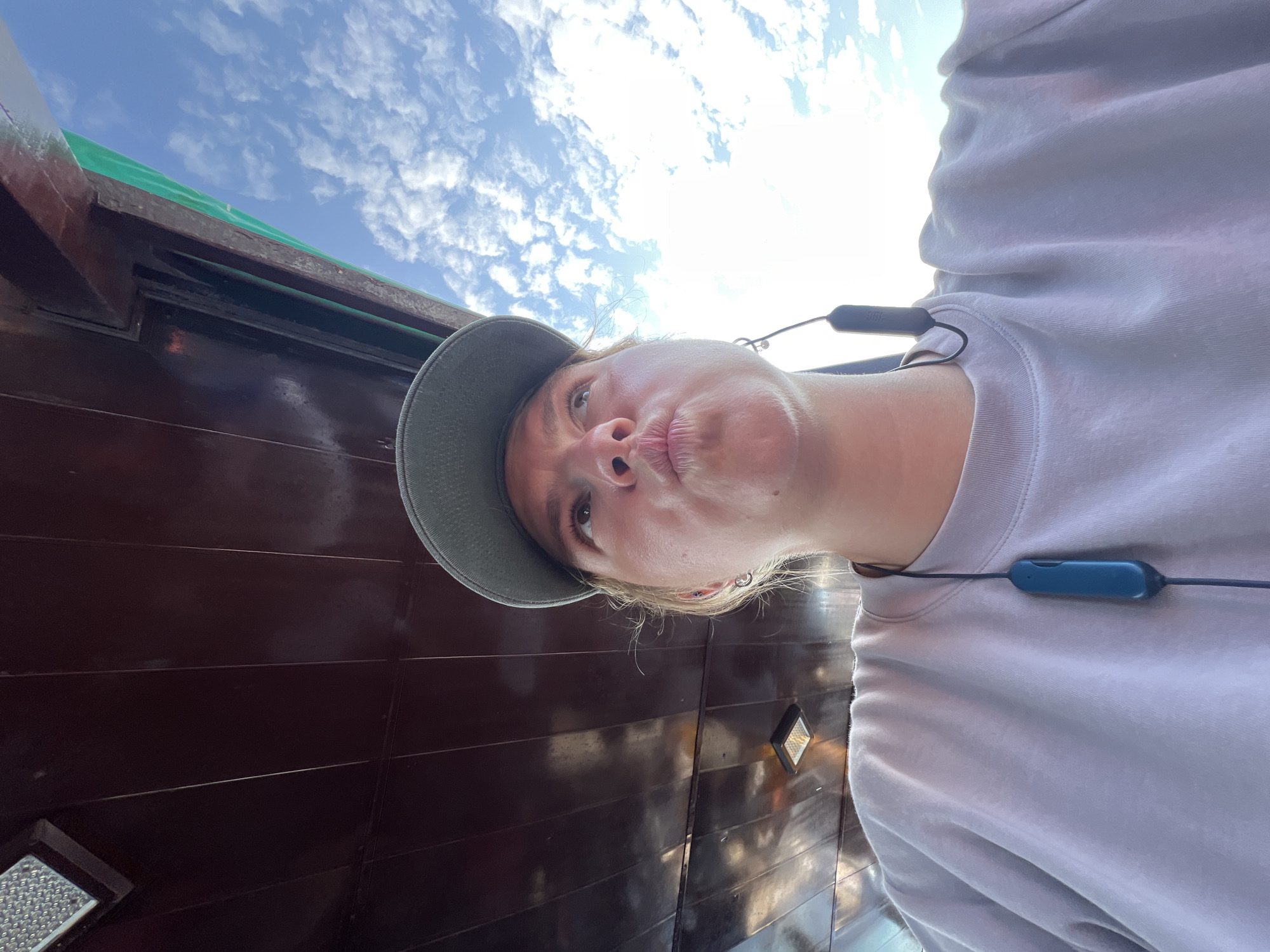 A person wearing a gray sweatshirt and a gray cap takes a selfie looking upwards with pursed lips and a building behind them. The background shows a wooden structure with some green covering the roof, and a cloudy sky above. The location is likely Nha Trang Ward, Vietnam, given the prompt's explicit location information. The person also has earbuds around their neck.