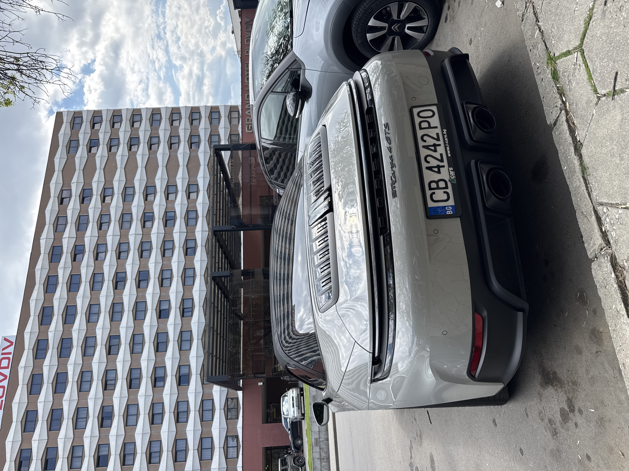 A light grey Porsche 911 Targa 4 GTS is prominently parked on a paved street, viewed from its rear-right quarter. The car's Bulgarian license plate, "CB 4242 PO," is clearly visible, along with the country code "BG" and the website "www.i-cars.bg." The model designation "PORSCHE 911 Targa 4 GTS" is emblazoned across the rear of the vehicle, which features quad black exhaust pipes. Parked directly in front of the Porsche is another grey car, possibly an SUV, partially visible, identifiable by its Michelin tires and a Citroën-like wheel hub.
In the background, a multi-story modern building with a distinctive facade stands tall. The facade features a repeating geometric pattern of light-colored, possibly white or cream, panels forming rhomboids, alternating with dark, recessed windows. The lower section of the building has a darker, reddish-brown tone. Visible text reflected in the Porsche's windows, and also directly on the building, indicates "GRAND HOTEL PLOVDIV." The word "PLOVDIV" is also visible in red lettering on a vertical banner-like structure attached to the building's side.
The scene takes place outdoors during the daytime under a partly cloudy sky. The ground consists of concrete paving, with some cracks, moss, and debris visible along the curb on the right. No people are present in the image. The setting is an urban environment, specifically in Plovdiv, Bulgaria, as indicated by the building name and license plate.