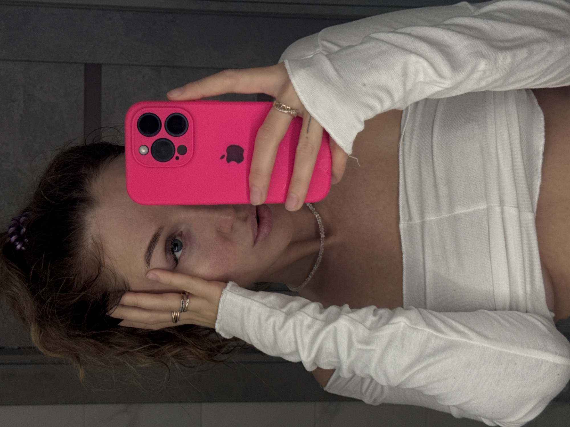 A person with light skin and blue eyes is holding a pink iPhone in front of their face in a selfie-style image. The phone has a black Apple logo and three camera lenses. The person is wearing a white, long-sleeved top and a delicate silver necklace. They have rings on both hands. Their hair is brown and slightly wavy. The background appears to be a grey wall, possibly in a bathroom or a room with similar decor. The location, Chaikovsky, Russia, is not discernible from the image.