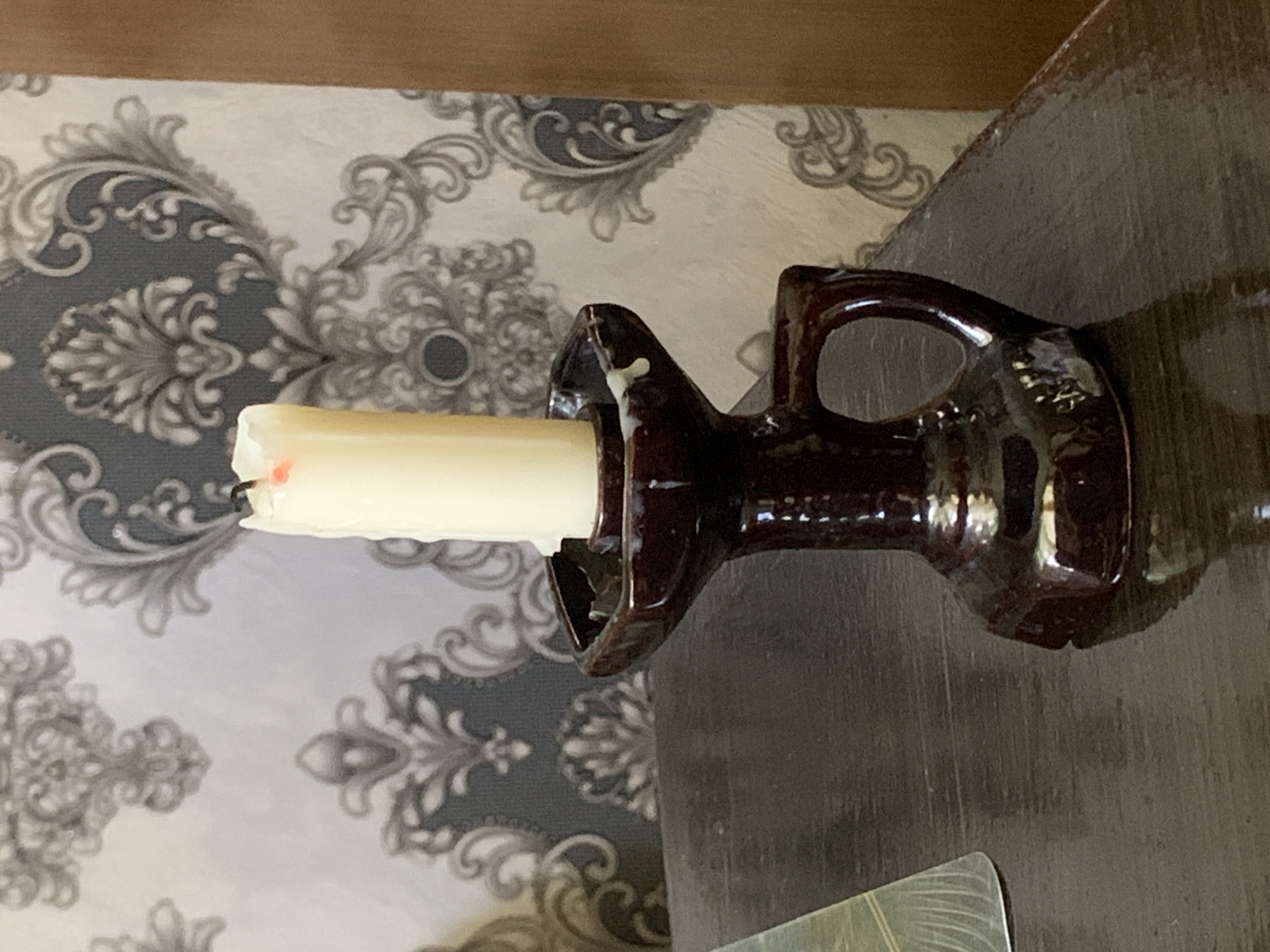 A single, unlit, cream-colored candle stands upright in a dark brown, ceramic candlestick holder. The candlestick has a flared base and a handle. The candle appears to have melted wax drippings. The candlestick is positioned on a dark, reflective wooden surface, likely a table. In the background, a wall is covered with ornate, patterned wallpaper in shades of gray and white, featuring floral and scroll motifs. To the right, a portion of a brown wooden door or furniture is visible. The scene is an interior setting. The image does not provide any specific location markers for Tashkent, Uzbekistan.