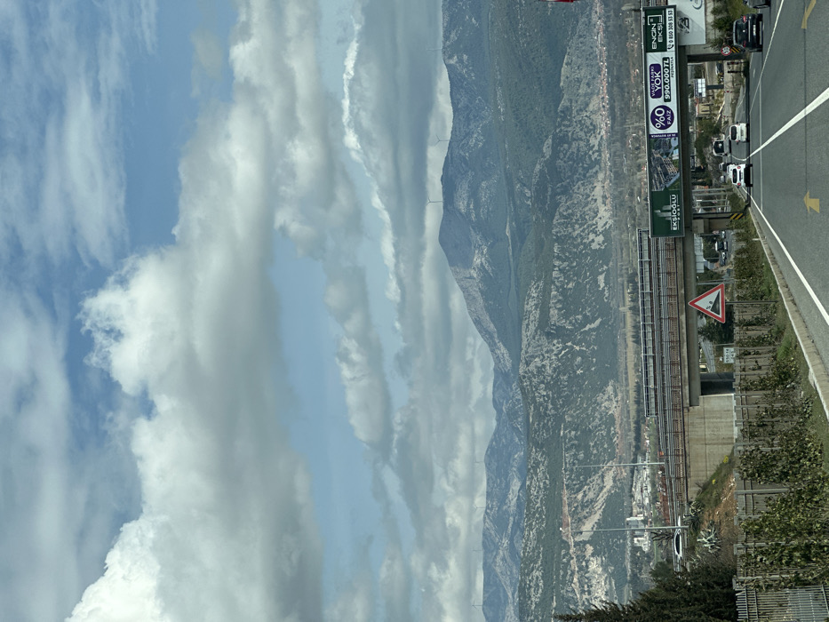 The image presents an outdoor scene depicting a multi-lane highway in the city of Menteşe, Turkey, under a partially cloudy sky. The highway, marked with white lines and a yellow directional arrow, extends towards the viewer and then under a concrete overpass. Multiple vehicles, primarily white cars and at least one white bus, are observed traveling away from the viewer on the highway.
In the background, a range of rocky mountains, partially covered with sparse green vegetation, dominates the horizon. Several wind turbines, approximately six, are visible atop or along the ridges of these mountains. On the lower slopes and in the valley between the mountains and the foreground, an urban development consistent with Menteşe is visible.
In the midground, a concrete overpass spans horizontally across the highway. Attached to this overpass are large green billboards displaying Turkish advertisements, including "EKŞİOĞLU," "%0 FAİZ," and "990.000 TL." To the right of the highway, before the overpass, a red and white triangular road sign indicating falling rocks is visible. The immediate left foreground features a dark green coniferous tree and a metal fence lined with green shrubs. No individuals are explicitly depicted in the scene.